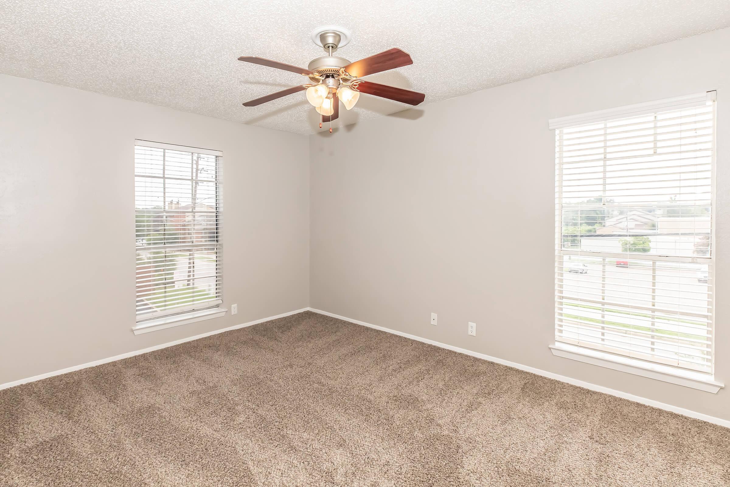 A vacant room featuring beige walls and plush carpet. Two windows with white blinds let in natural light. A ceiling fan with wooden blades and a light fixture is centered in the room. The space is empty, suggesting it is ready for new furniture or decorating.