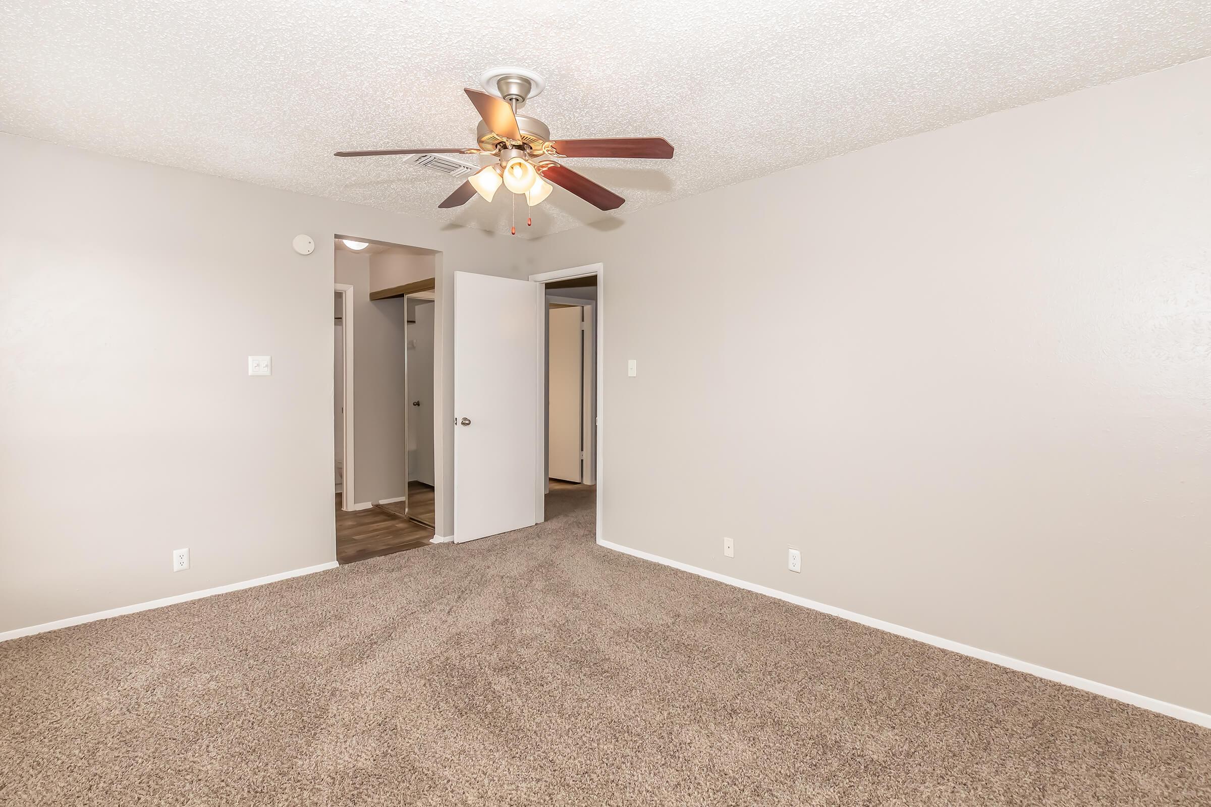 A spacious room with light beige walls and plush carpet, featuring a ceiling fan with wooden blades. A doorway leads to another room, and there is a closet visible on the left. Natural light enhances the inviting atmosphere.