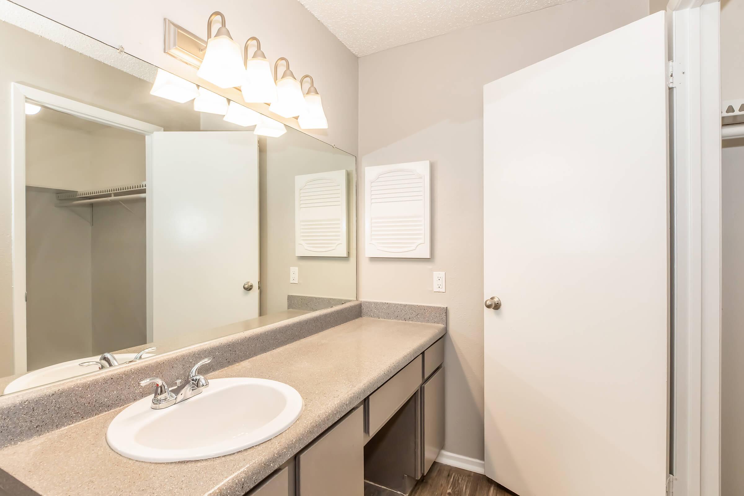 A modern bathroom featuring a double sink vanity with a gray countertop, a large mirror above the sink, and ample lighting. The walls are painted in a light color, and there is a closet with mirrored sliding doors on the left. Clean and minimalistic design with neutral tones.