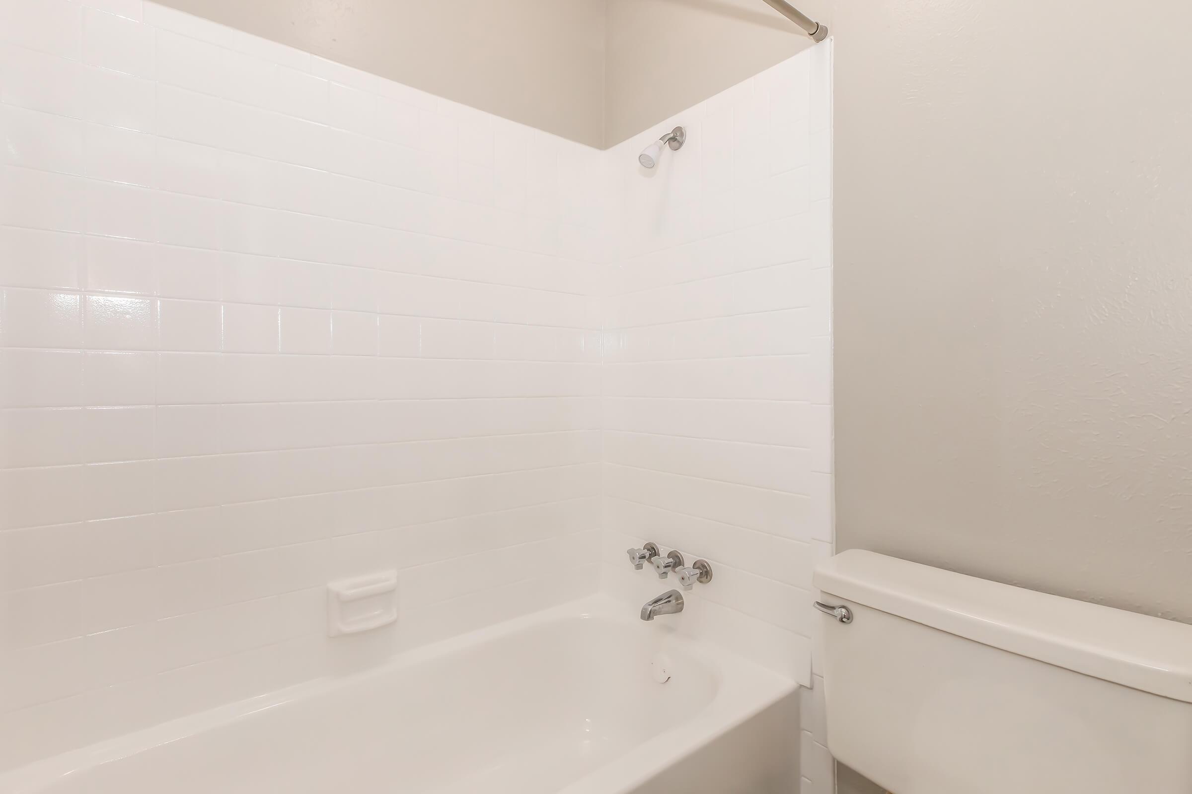 A clean, minimalist bathroom featuring a white tiled shower area with a bathtub, faucet, and showerhead. A white toilet is positioned next to the bathtub against a light-colored wall. The overall appearance is bright and uncluttered, emphasizing a simple and modern design.