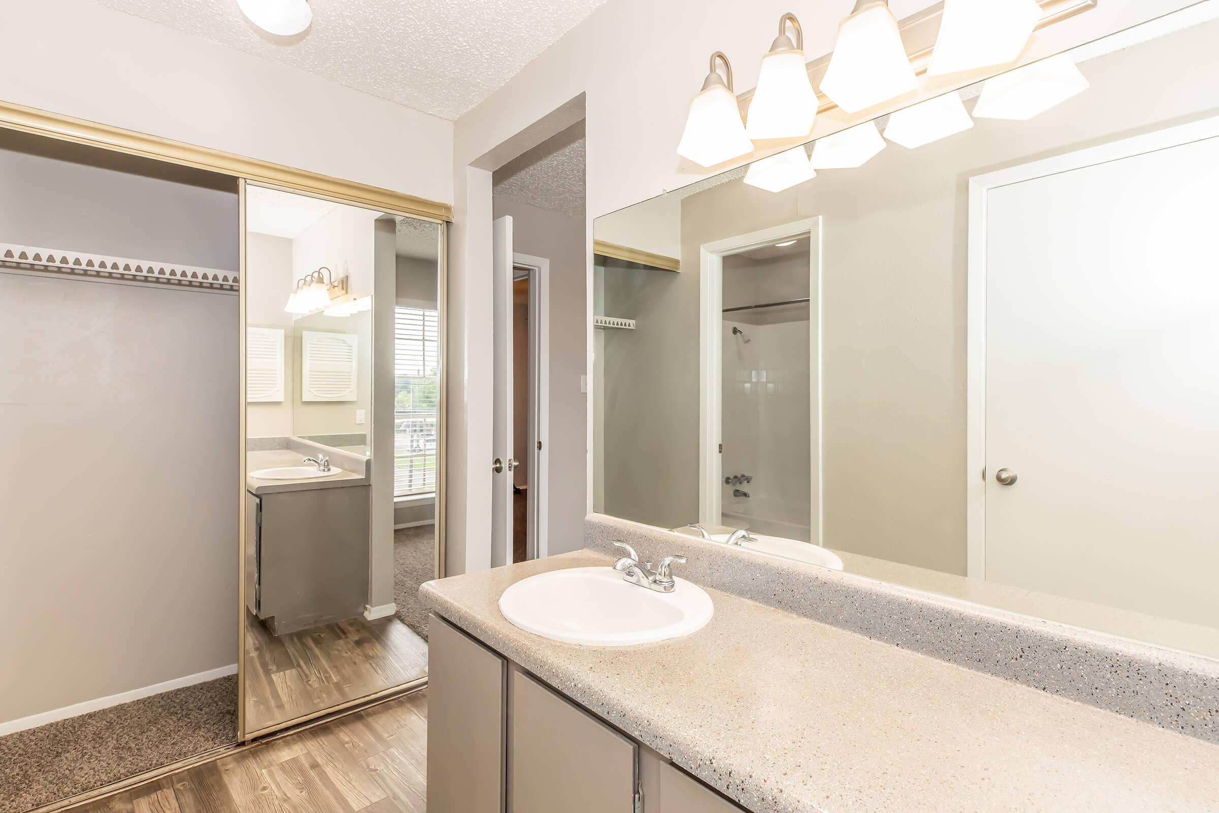 Modern bathroom featuring a double sink vanity with a light fixture above, a large mirror, and a mirrored closet door. The walls are painted a neutral color, and there is a window providing natural light. The flooring is a wood-like laminate, adding a warm touch to the space.
