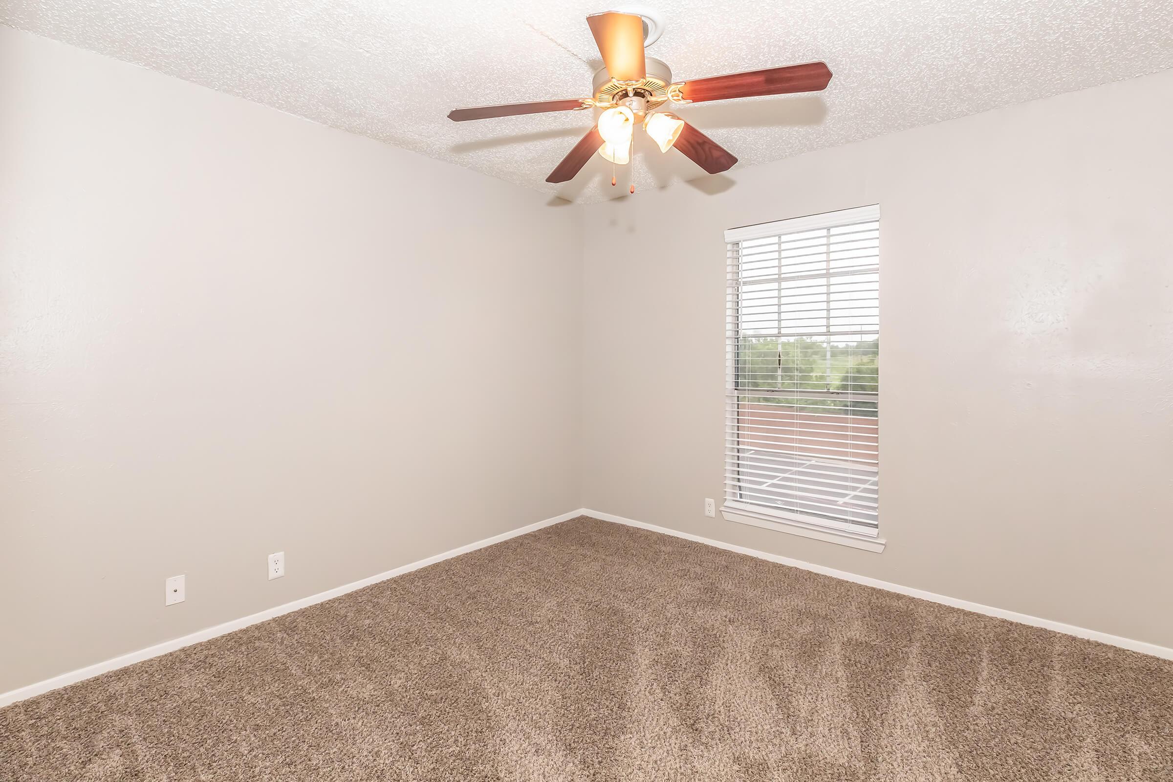 Empty room with beige walls and light brown carpeting. A ceiling fan with wooden blades hangs from the ceiling. There is a window with white blinds allowing natural light to enter. The space appears clean and well-maintained, suitable for various uses.
