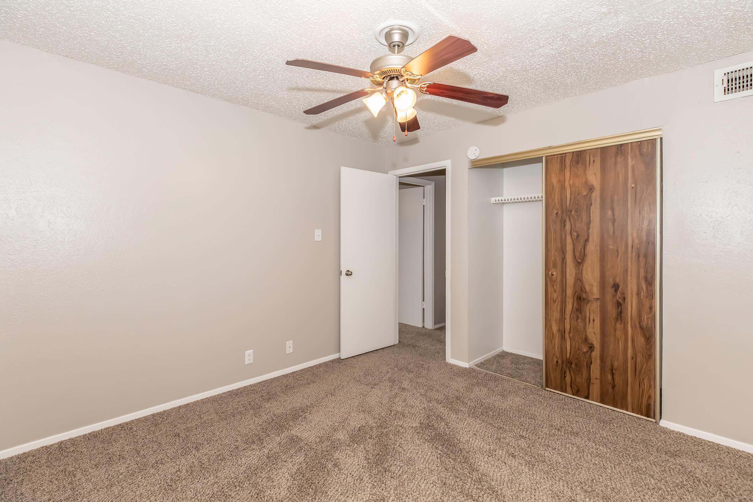A cozy bedroom with beige walls and light brown carpet. The ceiling features a fan with three lights. An open door leads to a closet with sliding wooden doors, while another door is visible, indicating access to another area. The room appears well-lit and spacious.