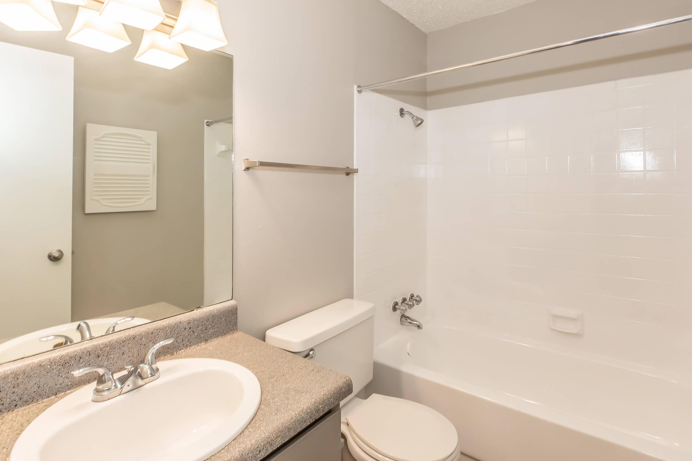A clean, well-lit bathroom featuring a white bathtub with a shower, a round sink with a faucet, a toilet, and a mirror above the countertop. The walls are painted in a soft gray color, and there are four light fixtures above the mirror, providing ample illumination.