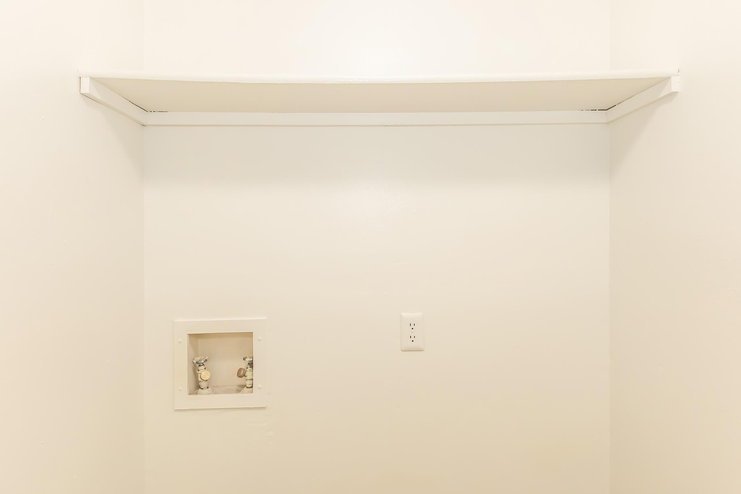 A minimalist empty room with white walls and a shelf mounted on the top. There is a small inset area on the wall with decorative objects, and an electrical outlet is visible below it. The overall atmosphere is clean and uncluttered.