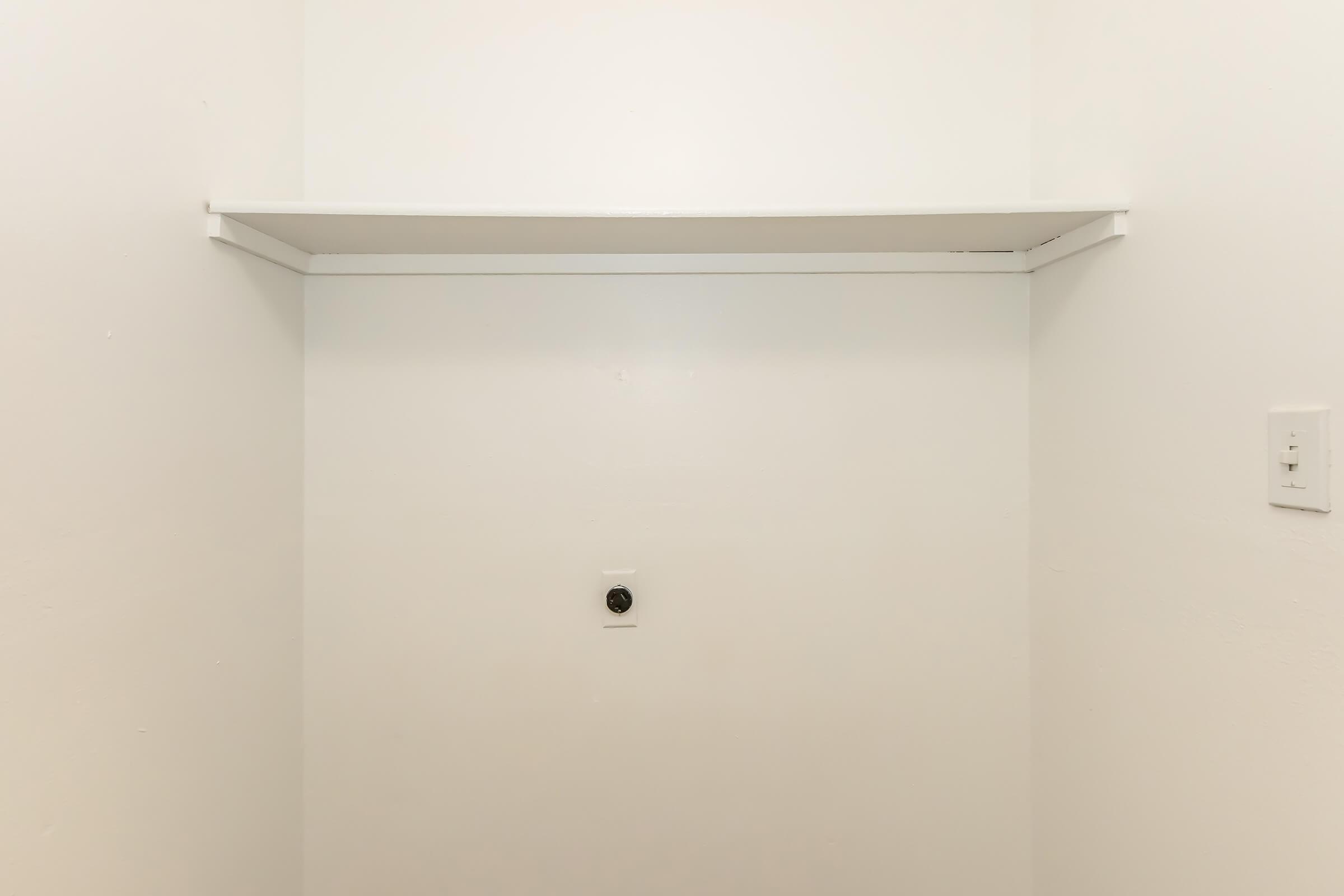 A clean, empty corner space with a single white shelf mounted against a plain white wall. There is a small electrical outlet below the shelf, and the overall feel is minimalistic and uncluttered.