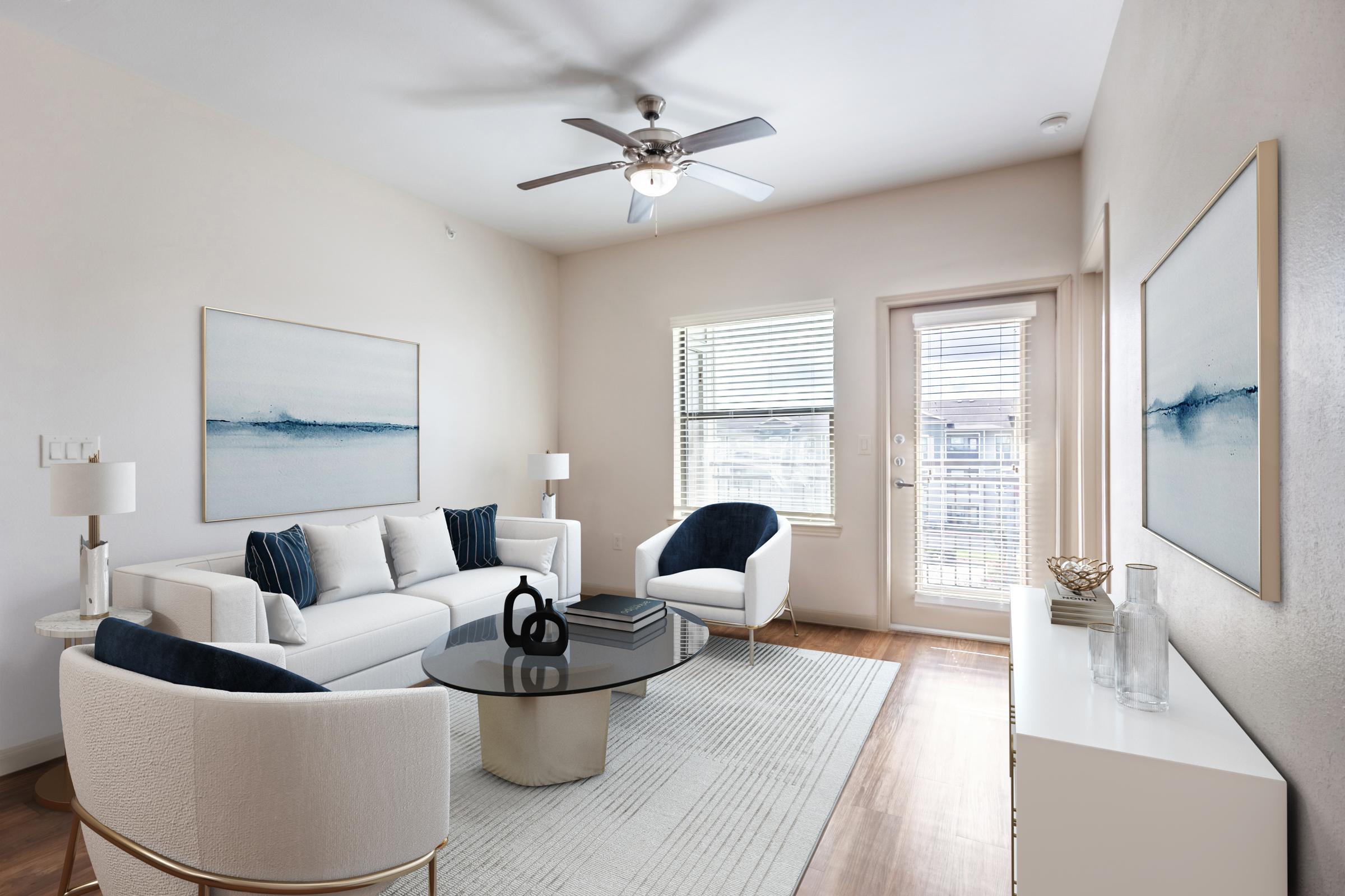A modern living room featuring a white sofa with blue accent pillows, two stylish armchairs, a round glass coffee table, and light-colored walls. Large windows provide natural light, and minimalist artwork hangs on the walls. A door leads to an outdoor space, enhancing the airy feel of the room.