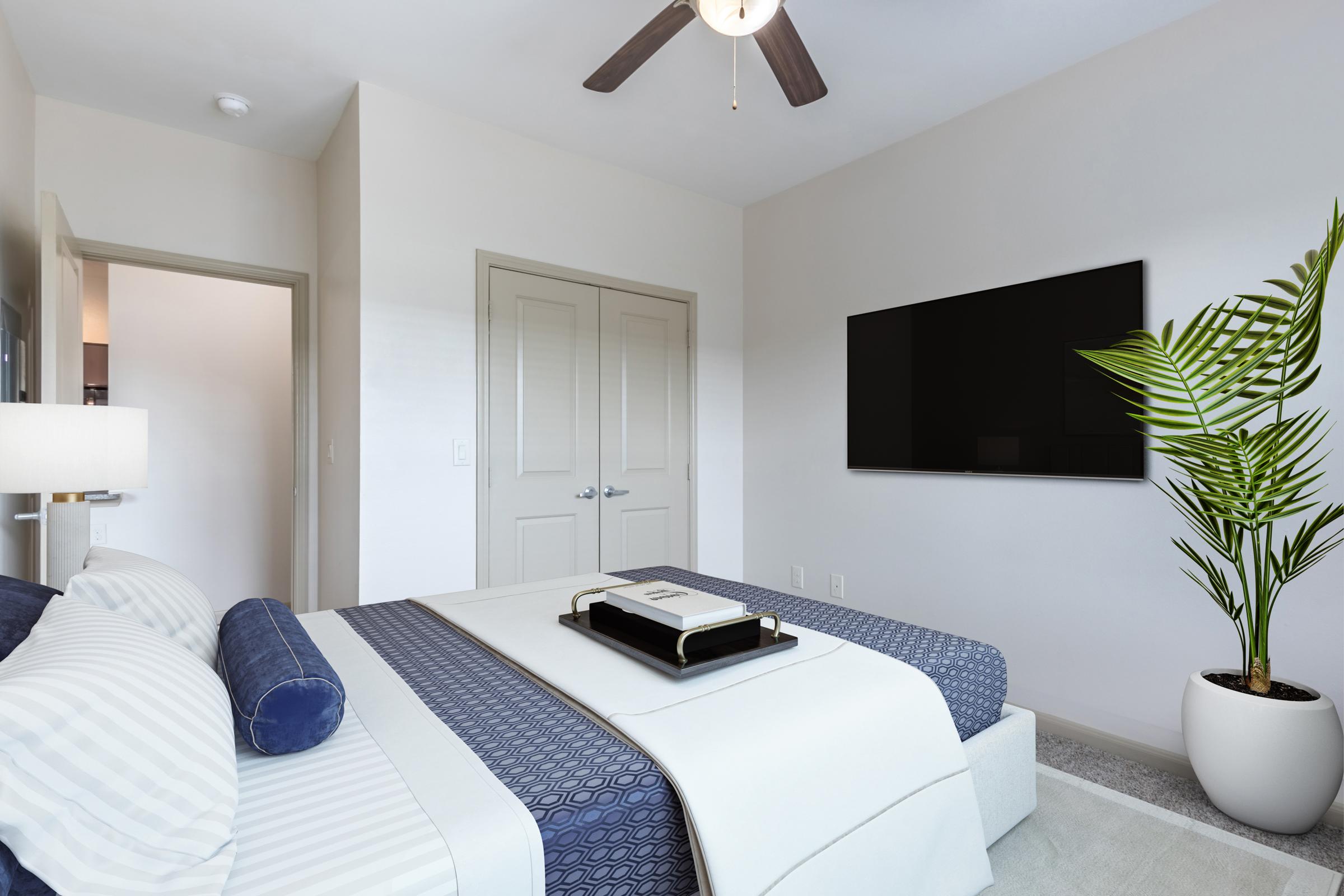 A modern bedroom featuring a neatly made bed with blue and white bedding, a decorative pillow, a plant in a white pot, and a wall-mounted flat-screen TV. The room has a neutral color palette, a ceiling fan, and a closed door leading to an adjoining space.
