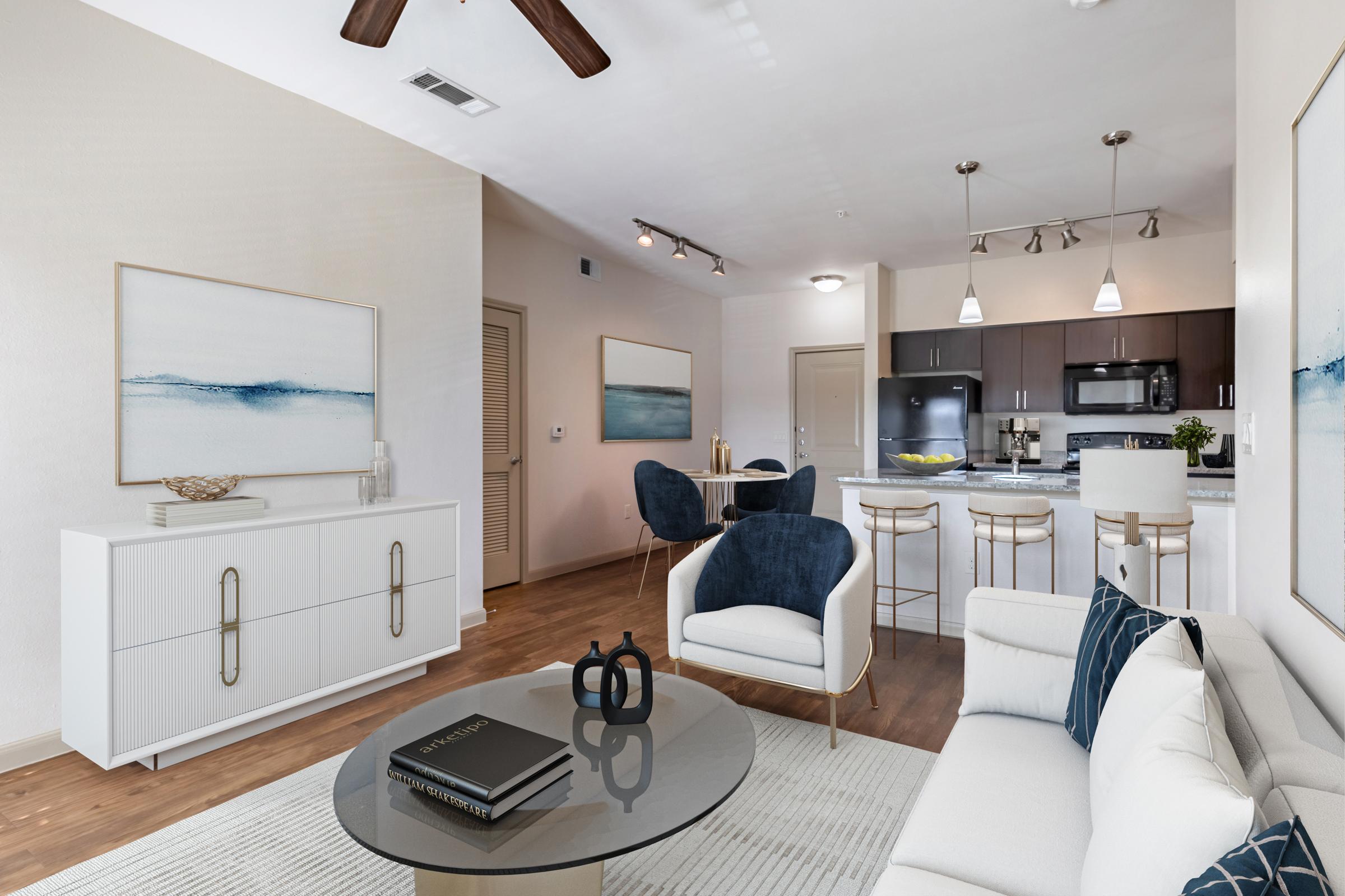 A modern living room featuring a white sofa and a glass coffee table with a decorative book. The space includes a dining area with dark chairs and a round table, a minimalist sideboard, and wall art depicting blue ocean waves. The kitchen behind has dark cabinetry and stainless appliances, creating a stylish, inviting atmosphere.
