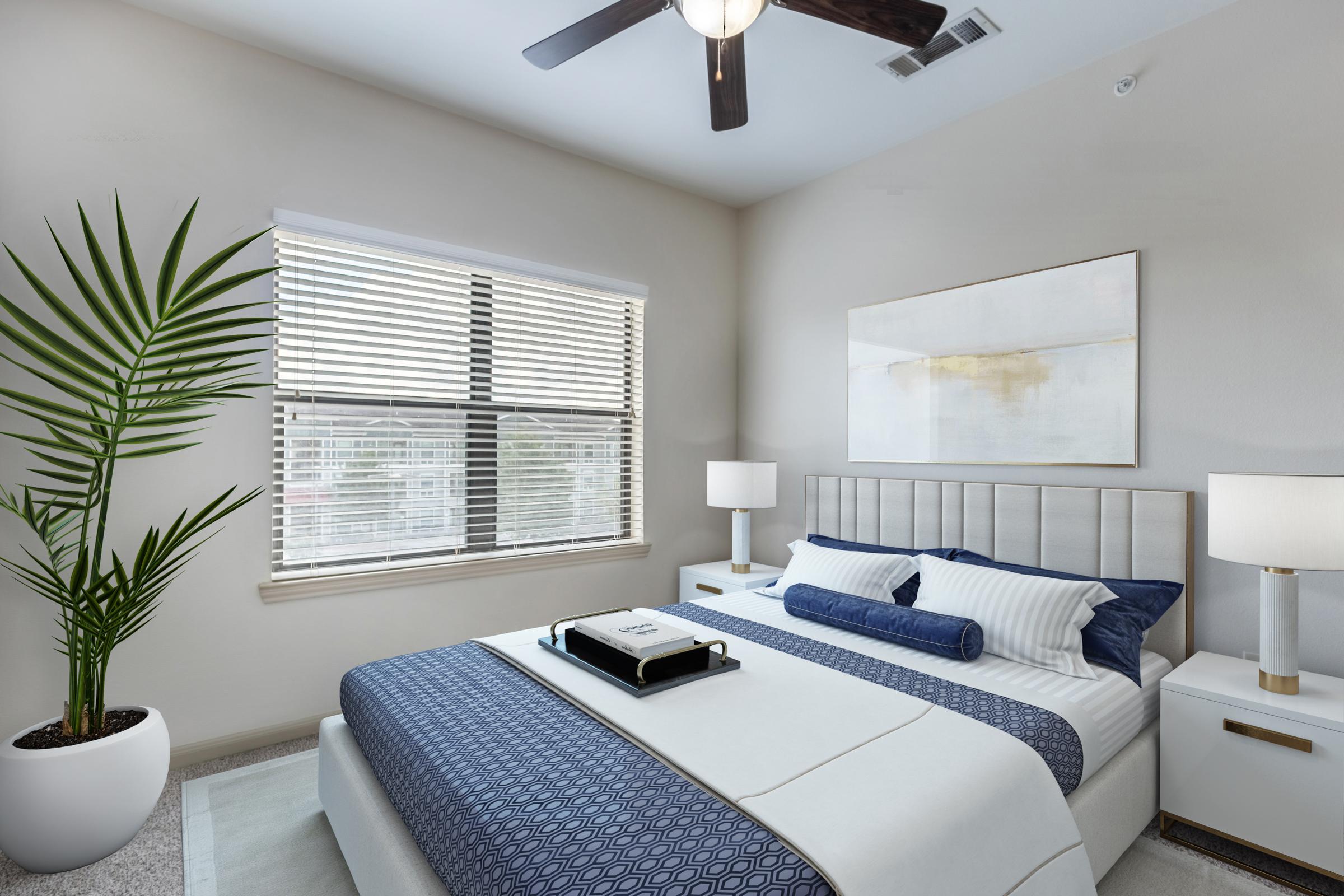 A modern bedroom featuring a light-colored wall, a neatly made bed with blue and white bedding, a decorative pillow, and two bedside tables with lamps. A large window allows natural light to flood the room, and a potted plant adds a touch of greenery to the space. A ceiling fan is visible above.