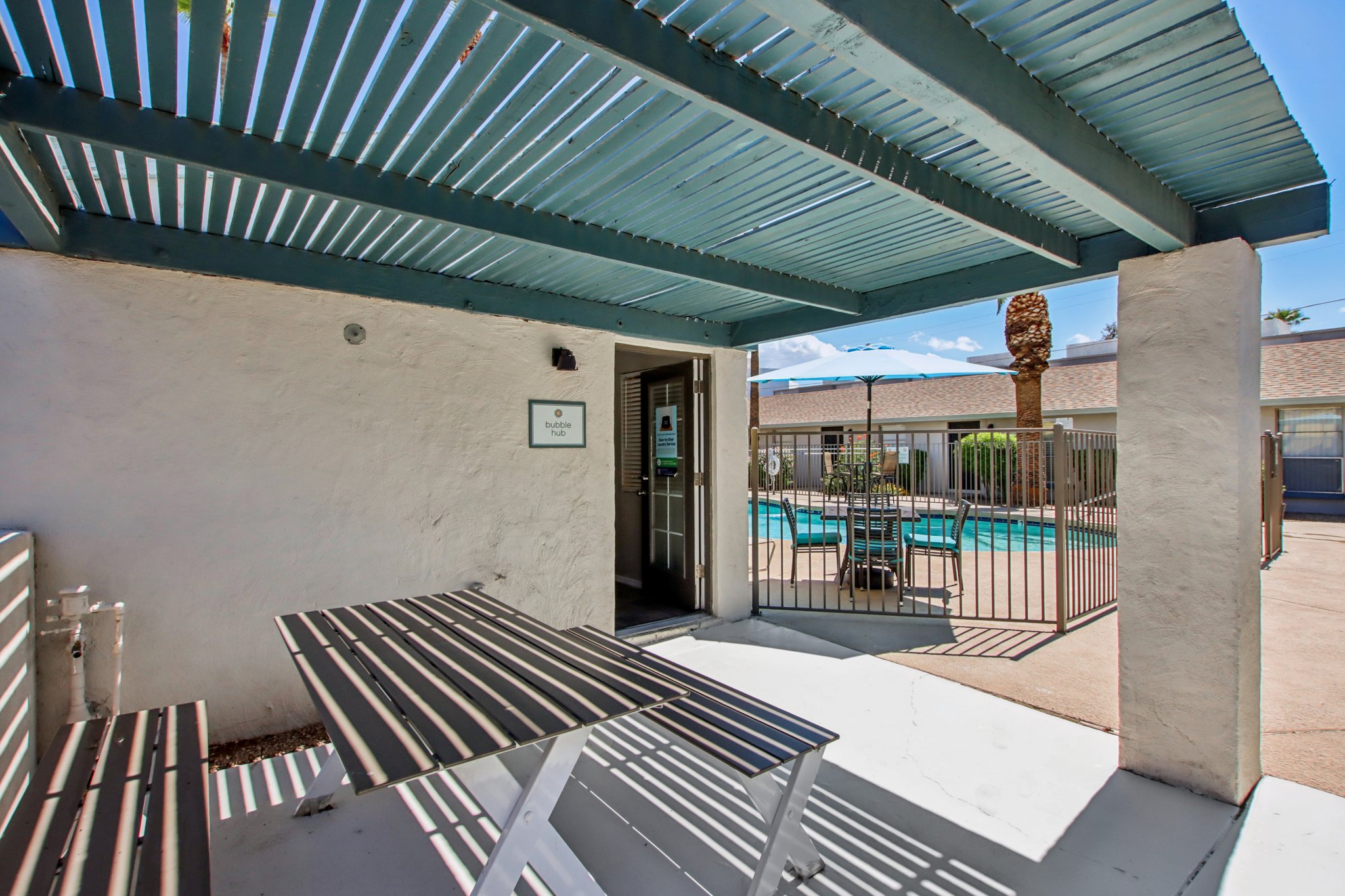 A shaded outdoor area with a picnic table and a view of a swimming pool beyond a gated fence. The structure has a white wall and blue overhead slats, while the sunny sky and palm trees are visible in the background.