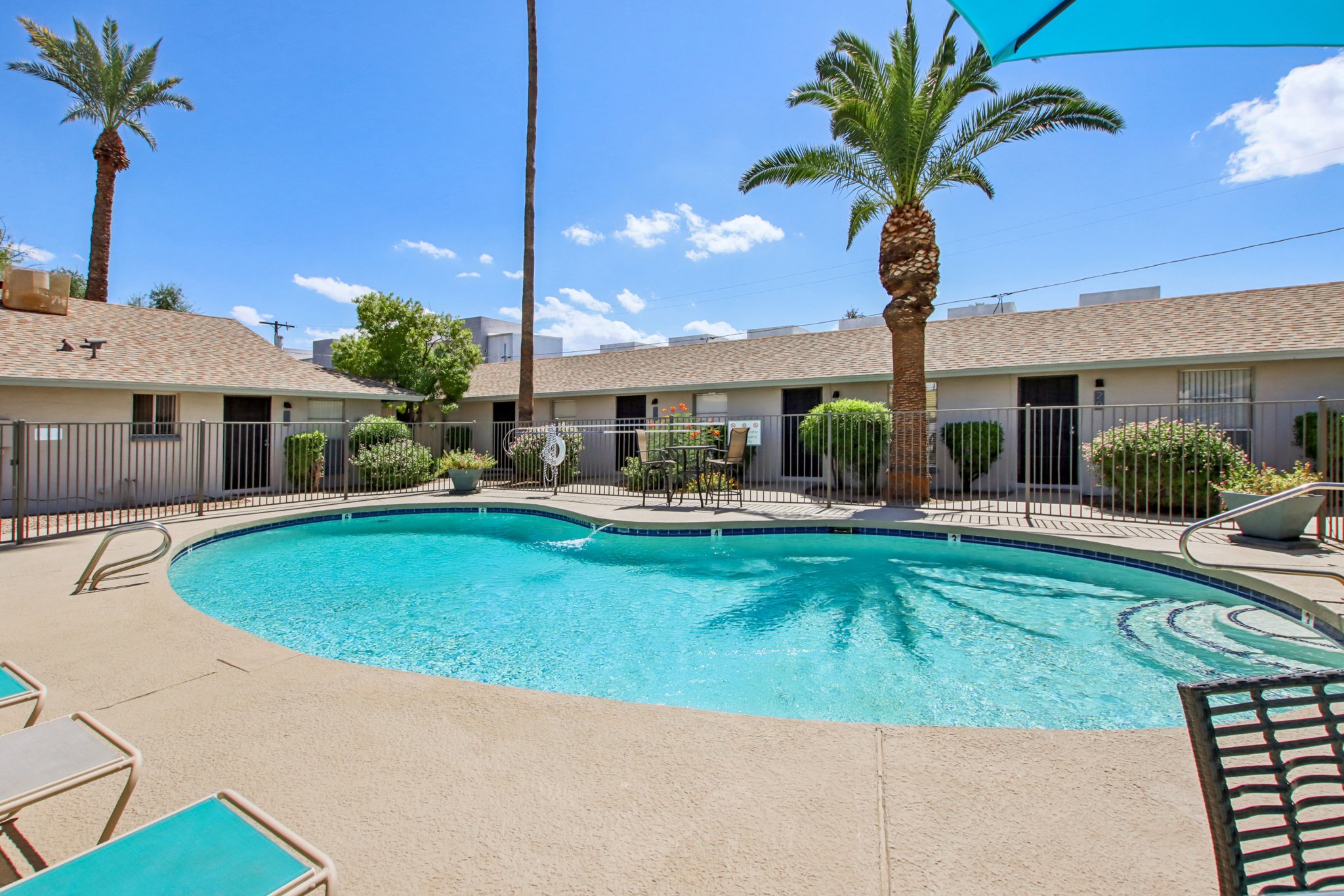 A bright outdoor swimming pool surrounded by palm trees and lounge chairs, with a small seating area nearby. The pool area is enclosed by a black fence, and there are buildings in the background under a clear blue sky dotted with a few clouds.