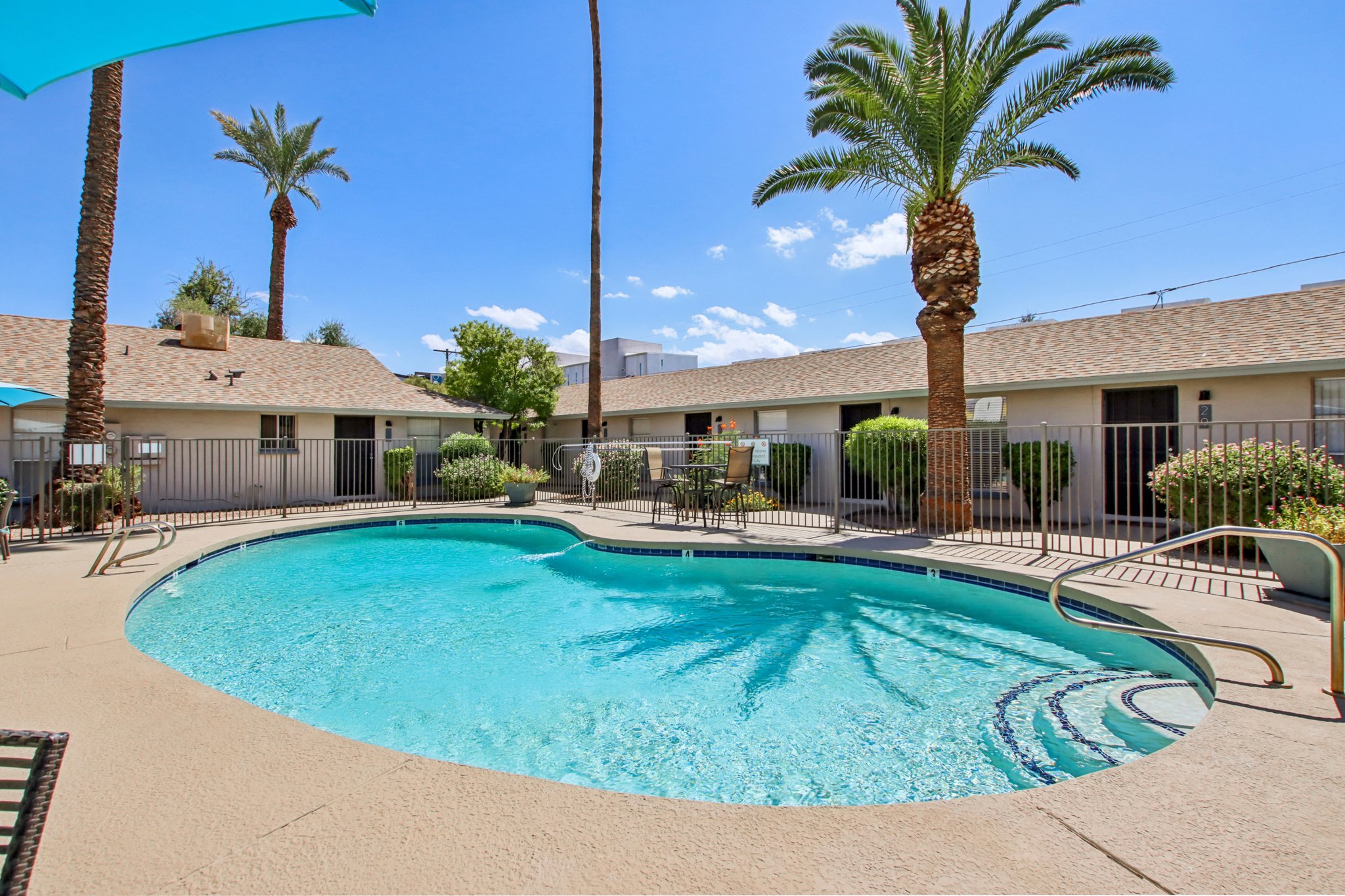 A bright outdoor swimming pool surrounded by palm trees, sun loungers, and patio furniture. The pool area features a fence and is located near buildings with a sunny sky and a few clouds in the background.