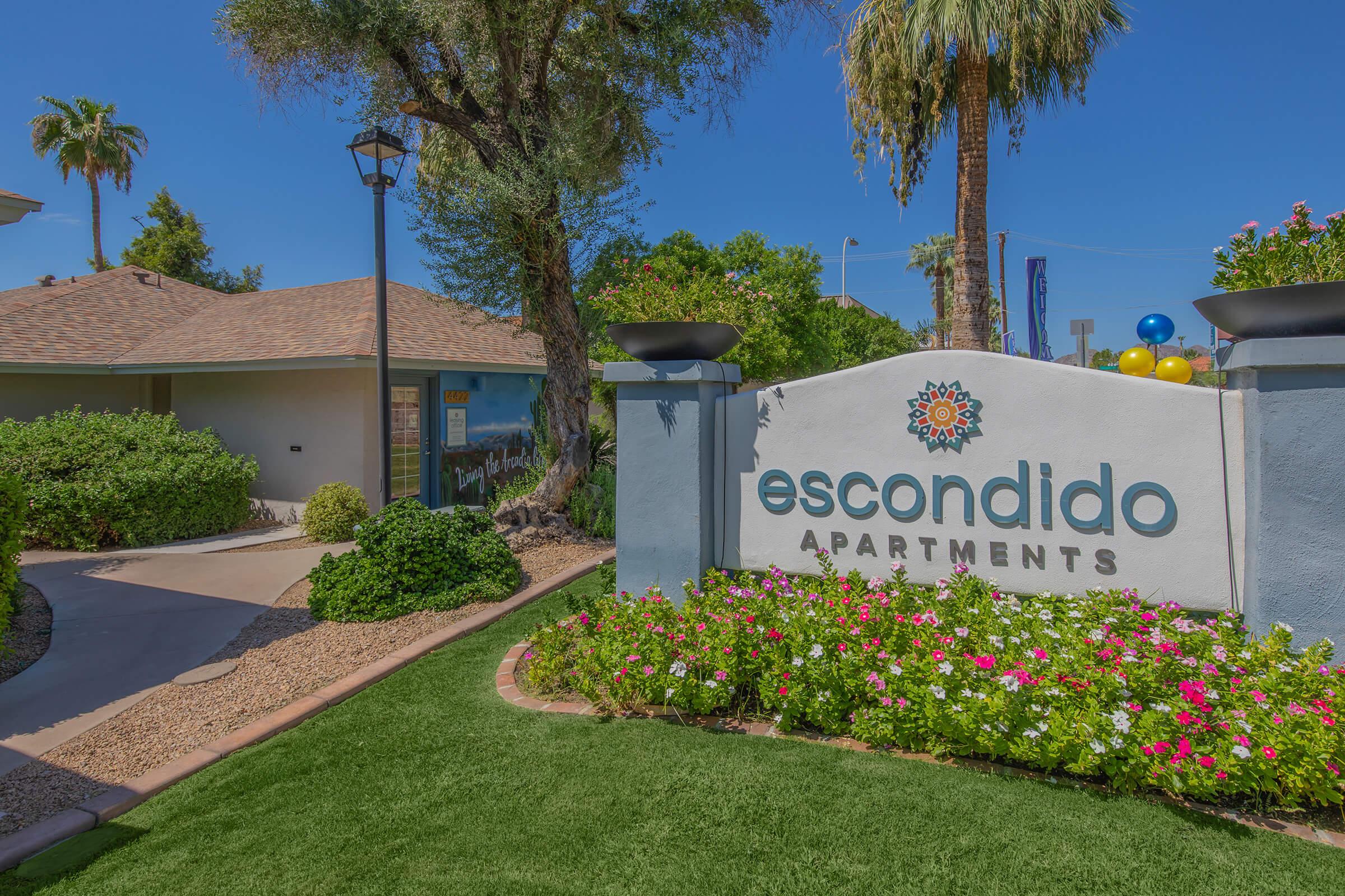 A welcoming entrance to Escondido Apartments featuring a large, decorative sign surrounded by vibrant flowers and lush greenery. The scene includes a residential building with a sloped roof and palm trees, set against a clear blue sky.