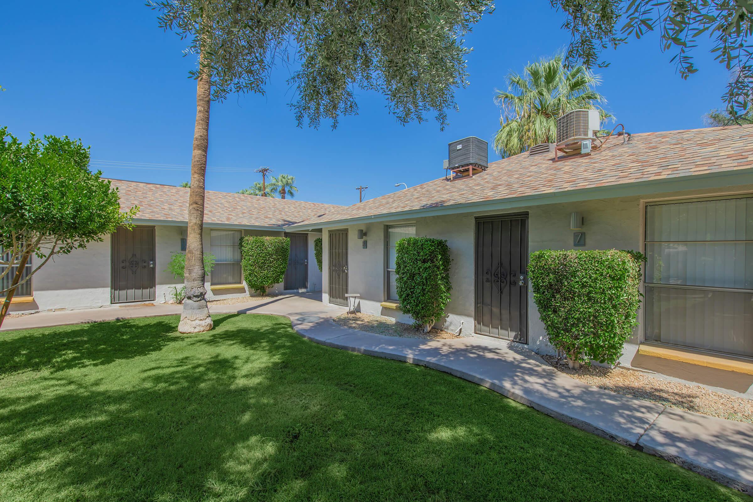 A sunny courtyard with well-maintained green grass and neatly trimmed shrubs. Several doors lead into a series of single-story buildings with a tiled roof. Palm trees and other landscaping provide a tropical feel, creating a welcoming outdoor space.