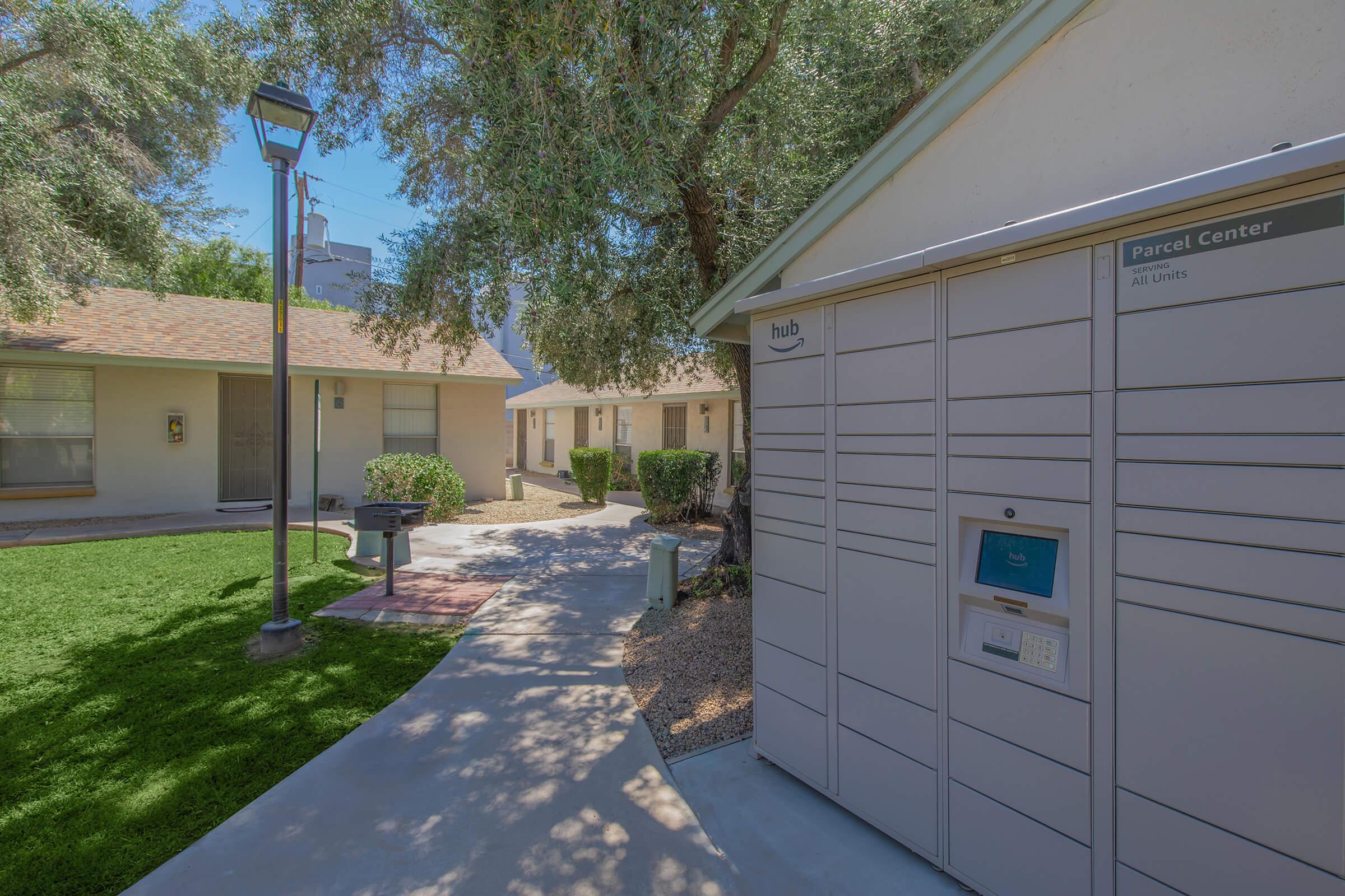 A pathway leading through a residential area with a parcel locker on the right. In the background, there are small houses and trees, providing a green, inviting atmosphere. A lamp post can be seen nearby, enhancing the ambient lighting.