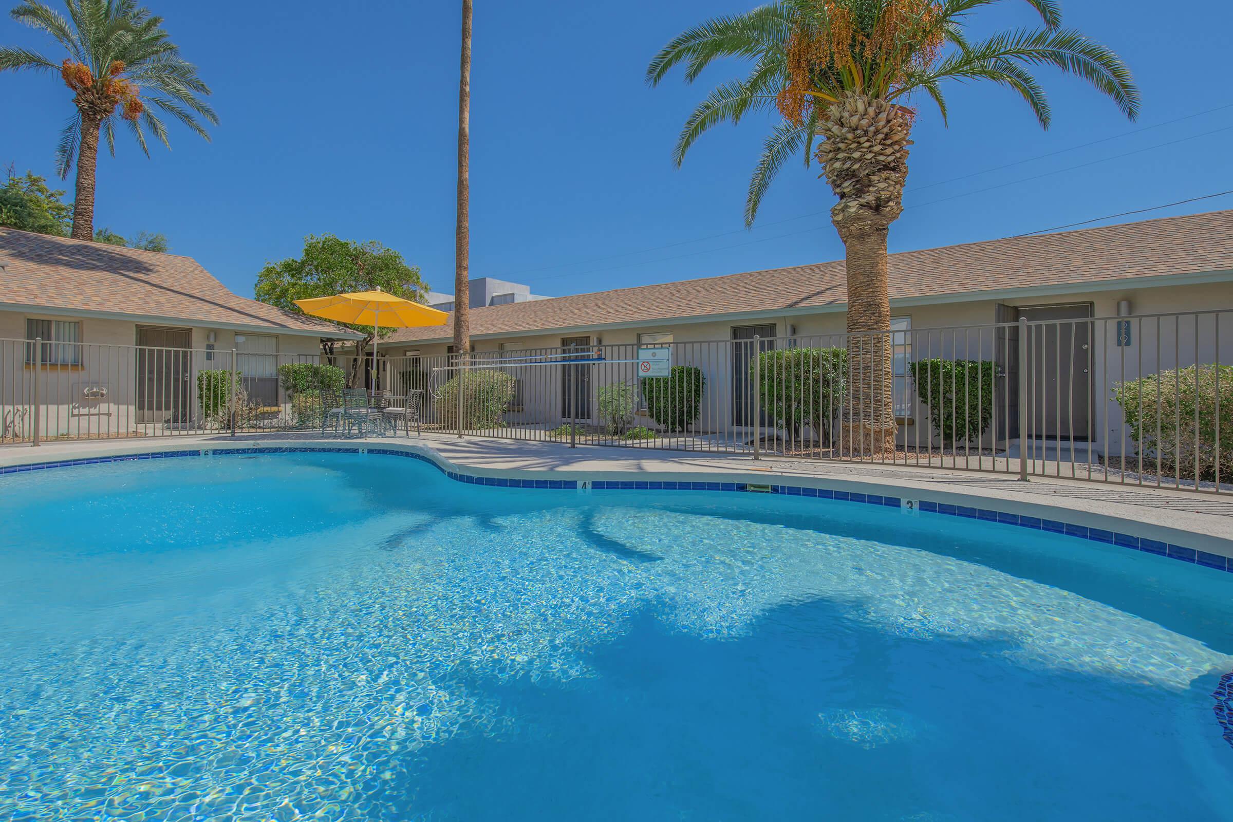 A clear blue swimming pool surrounded by palm trees and lush greenery, with lounge chairs and an umbrella. Nearby, there are several units with a beige exterior, creating a relaxed, vacation-like atmosphere. The sky is bright blue, indicating a sunny day.