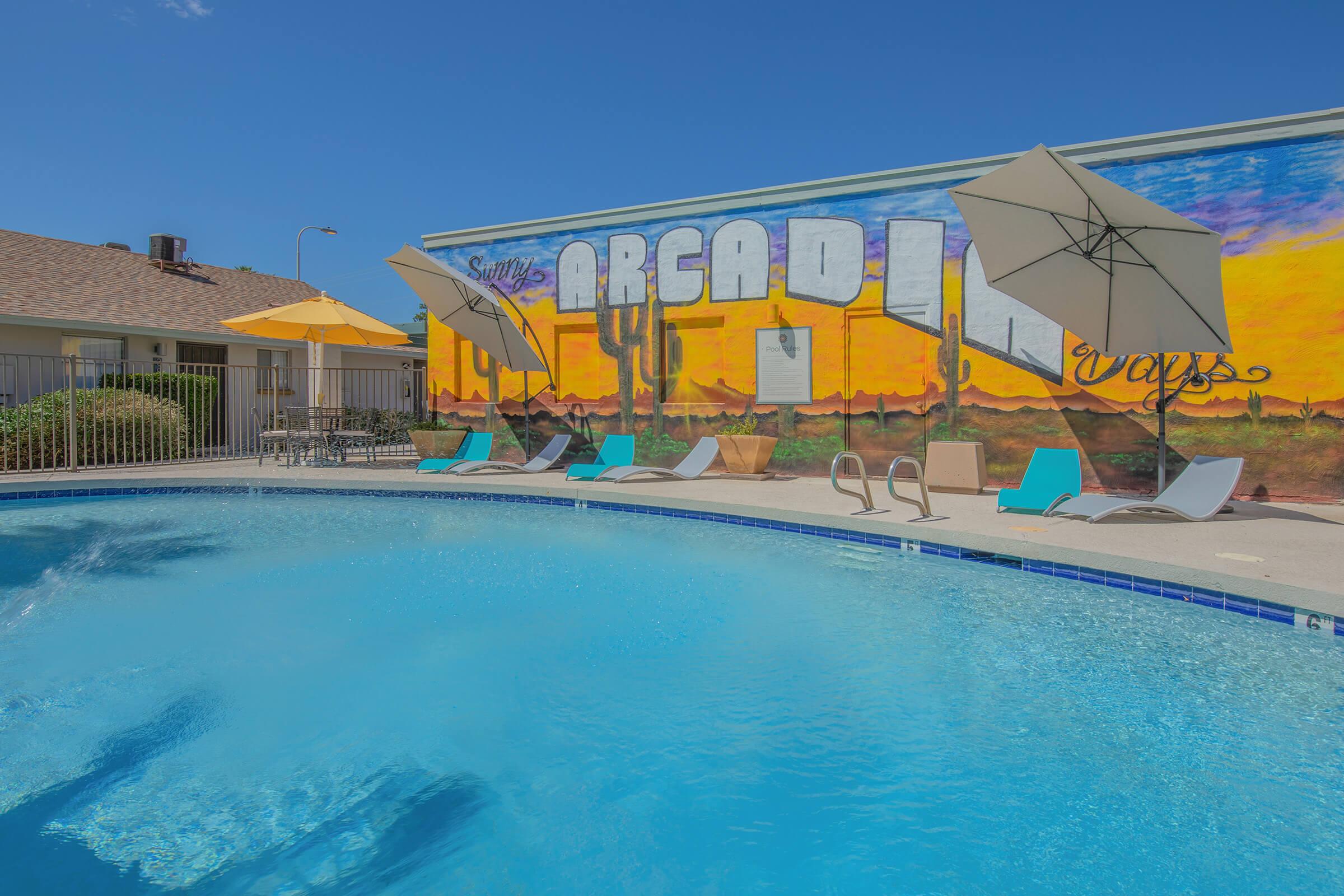 A bright blue swimming pool with lounge chairs and umbrellas on the side, set against a colorful mural of a desert landscape and cacti. The mural features bold text reading "Area" and "Days," enhancing the cheerful atmosphere of the area. Clear skies are visible above the scene.