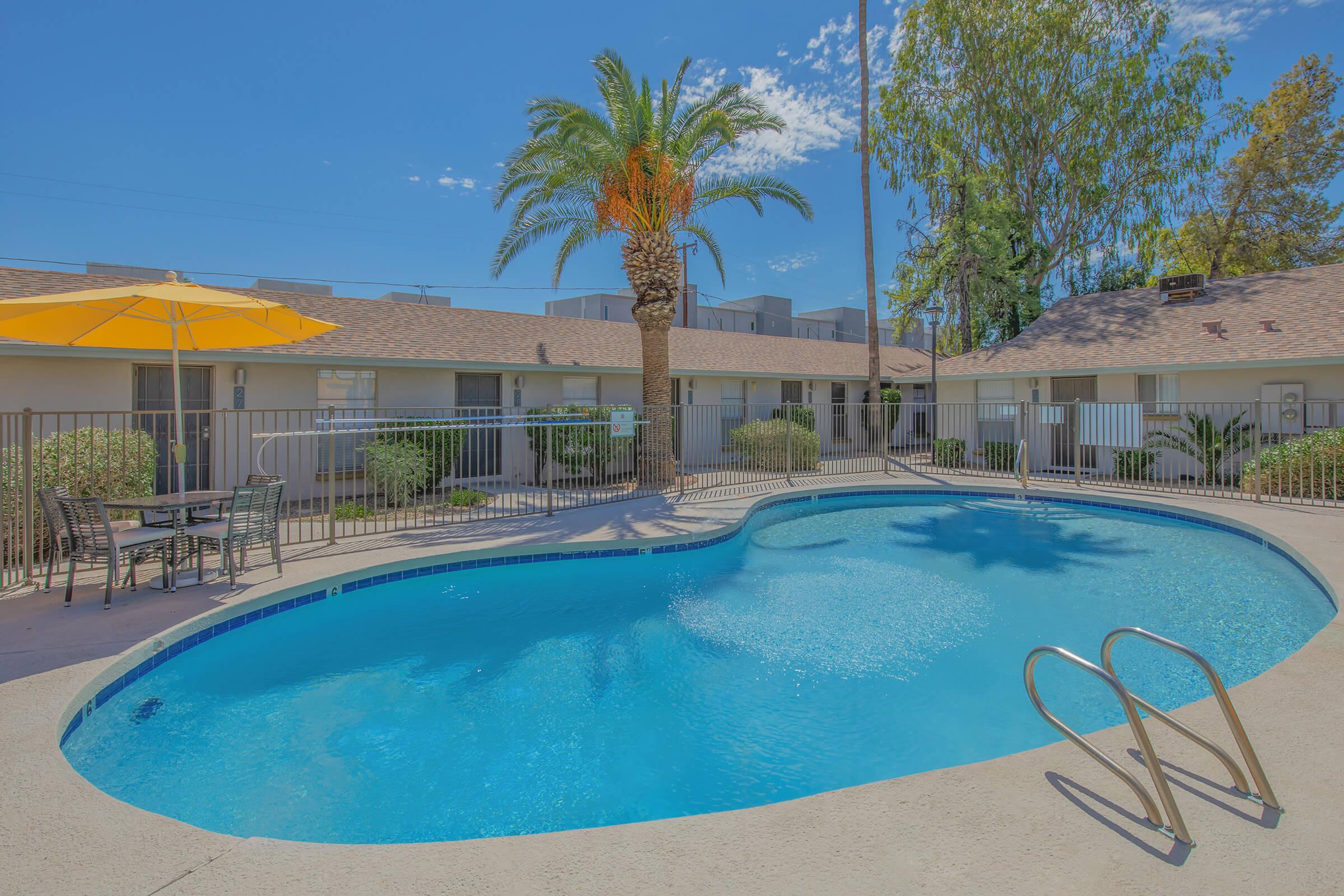 A sparkling blue swimming pool surrounded by palm trees and greenery. There are two lounge chairs and a table under an orange umbrella, with buildings visible in the background. The sky is clear and blue, adding to the relaxing atmosphere of the pool area.