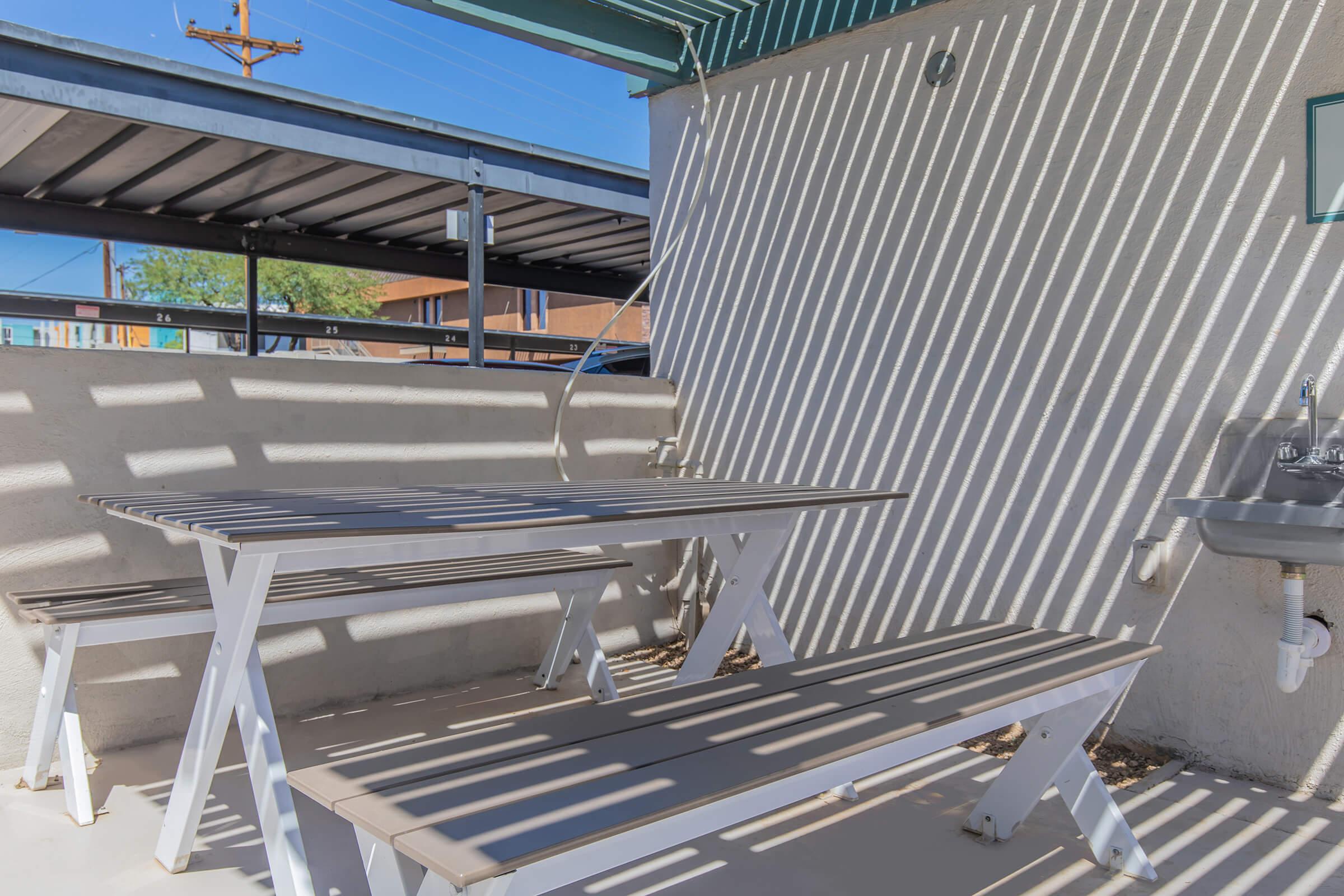 A shaded outdoor seating area featuring a white picnic table with benches under a slatted roof, with sunlight creating diagonal shadows on the surface. A small sink is visible on the wall nearby, surrounded by a concrete surface and a low wall. Background features a fence and buildings under a clear blue sky.