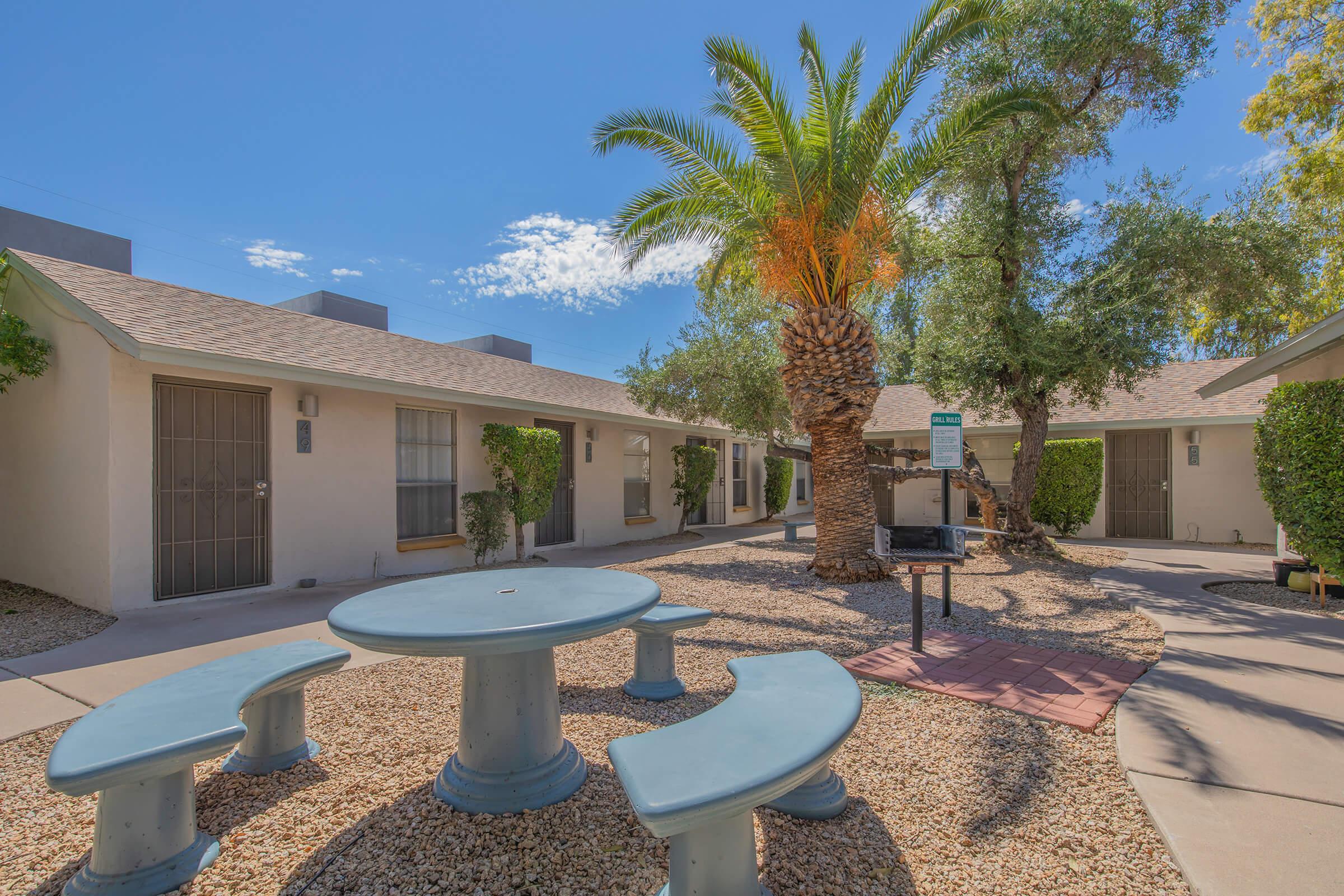 A courtyard featuring a round table with attached benches surrounded by small buildings, palm trees, and shrubs. The clear blue sky adds to the inviting atmosphere of this outdoor space, designed for relaxation and social gatherings.