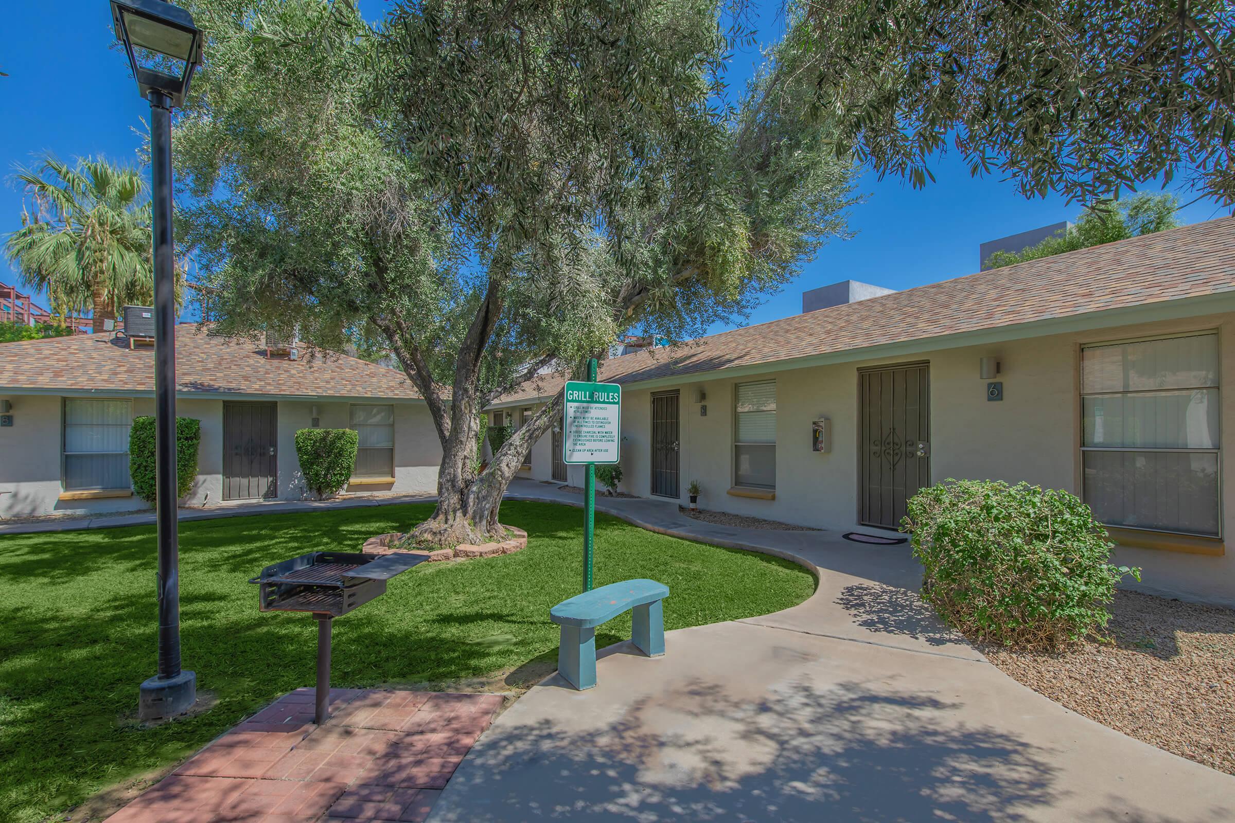 A well-maintained courtyard featuring several simple buildings with doors, framed by lush green grass and a tree. A blue bench is positioned near a sign, and there's a grill nearby. The setting is sunny, showcasing a peaceful and inviting outdoor space.