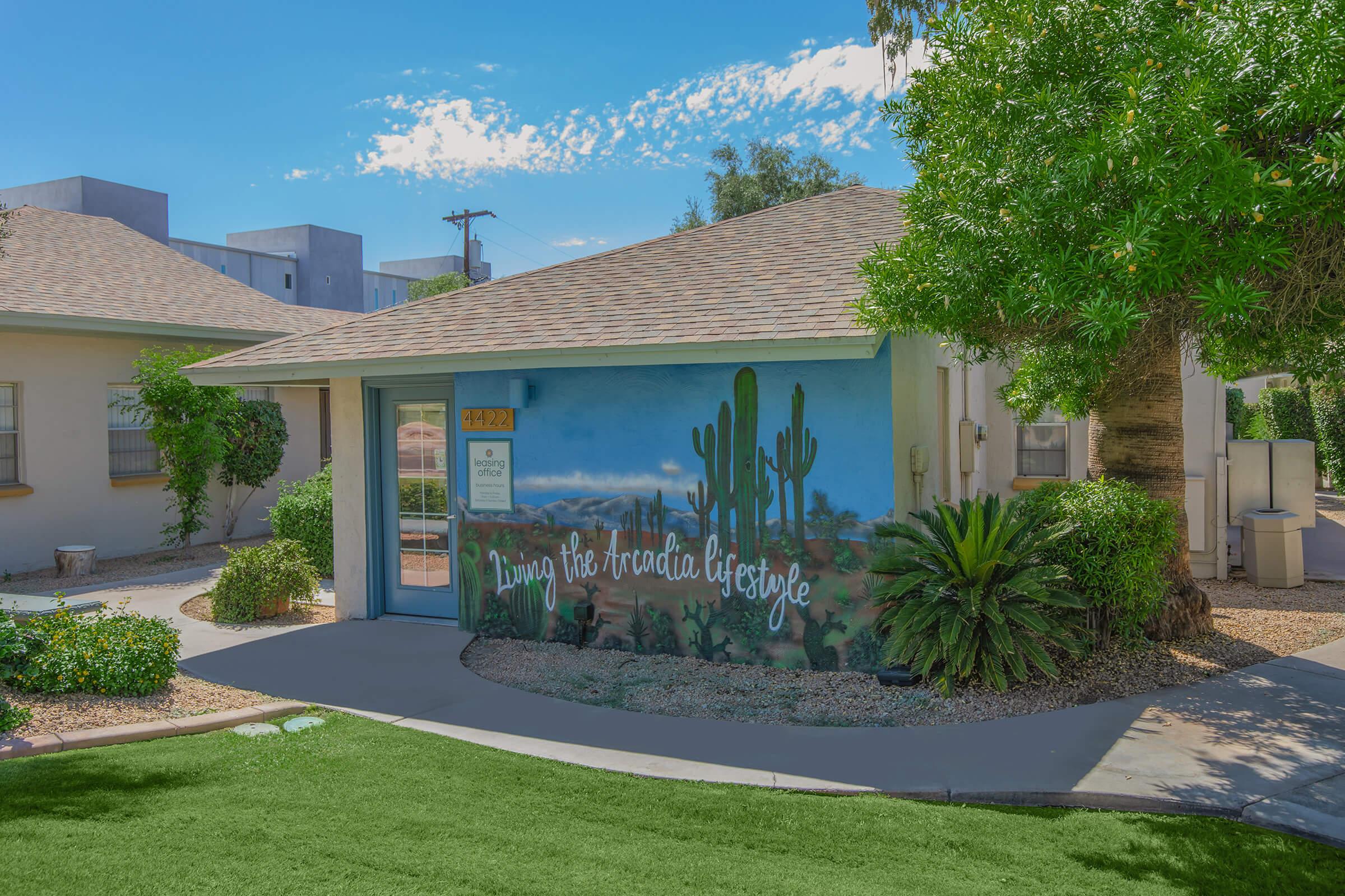 A view of a house with a colorful mural featuring cacti and the phrase "Living the Arizona lifestyle." The house has a sloped roof, surrounded by desert landscaping and green grass. In the background, a few other buildings are visible. The scene reflects a warm, sunny atmosphere typical of the Arizona desert.