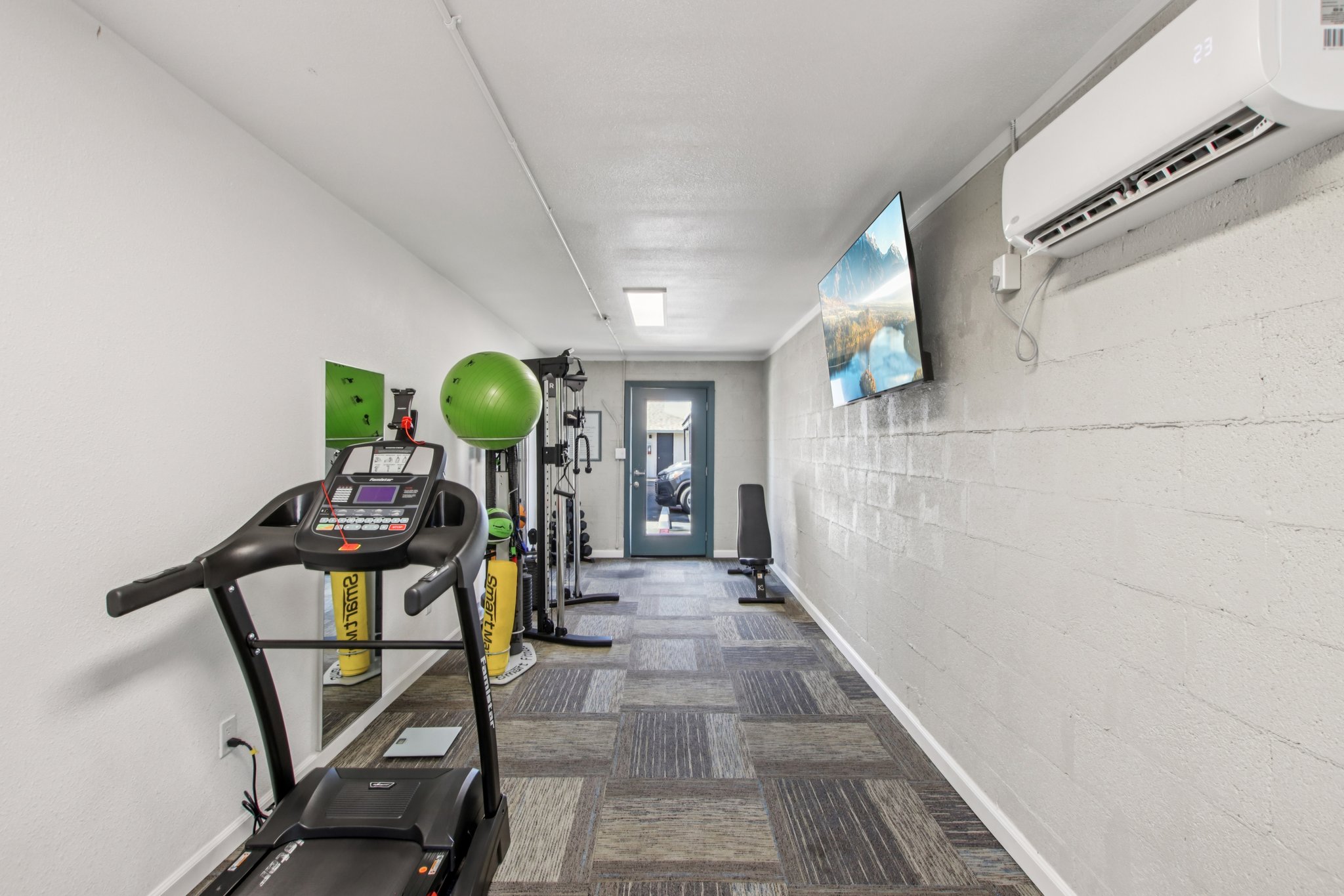 A view of a modern gym area featuring a treadmill, exercise balls, and fitness equipment against a clean, white wall. There's a large window with natural light and a television mounted on the wall. The flooring consists of gray and textured tiles, creating a welcoming workout environment.