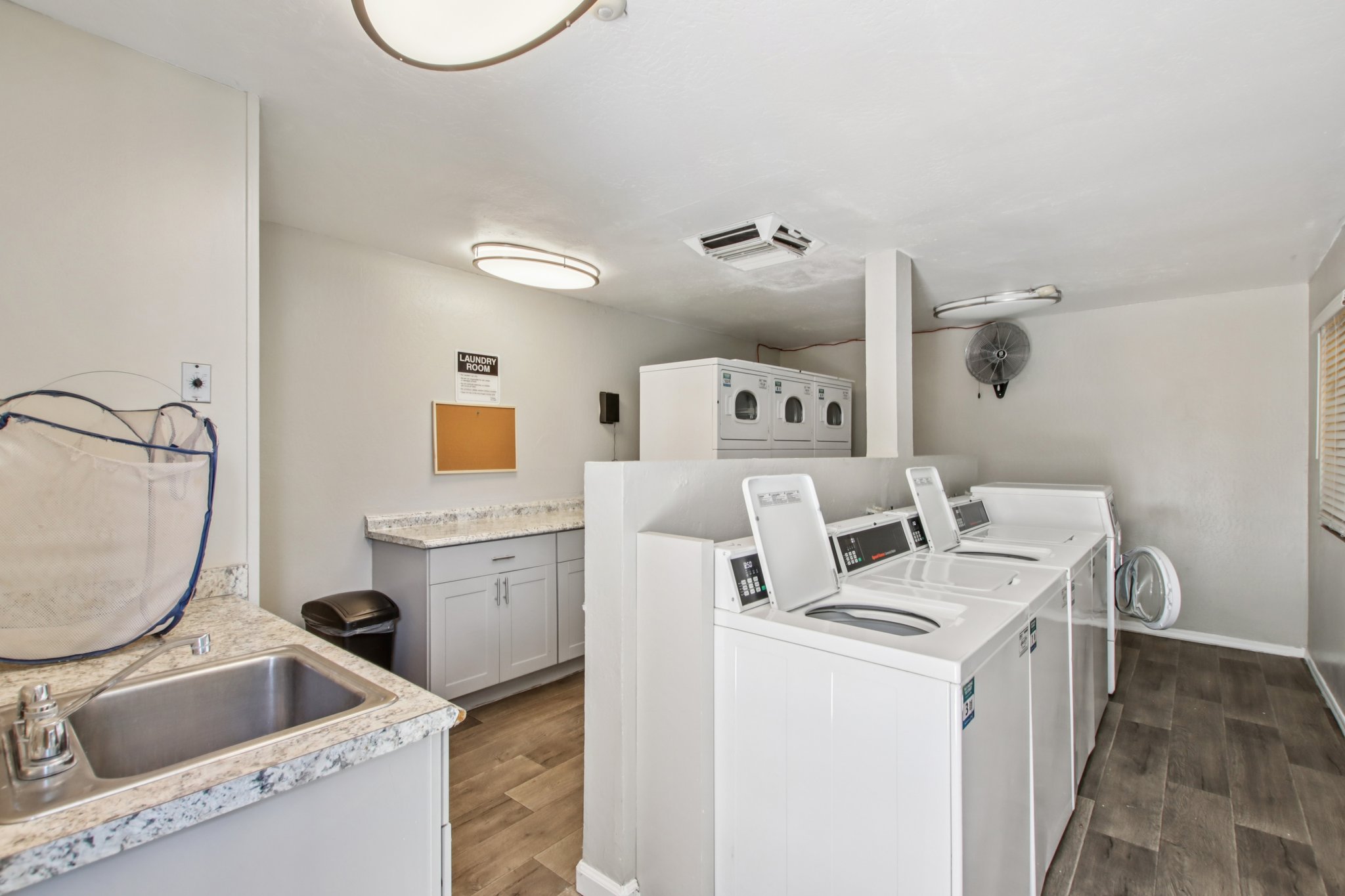 A laundry room featuring several white washing machines and dryers, a countertop with a sink, and a trash bin. The walls are light-colored, and there is a circular ceiling light and a wall-mounted fan. A bulletin board is visible on the wall. The floor is covered in light wood-like tiles.