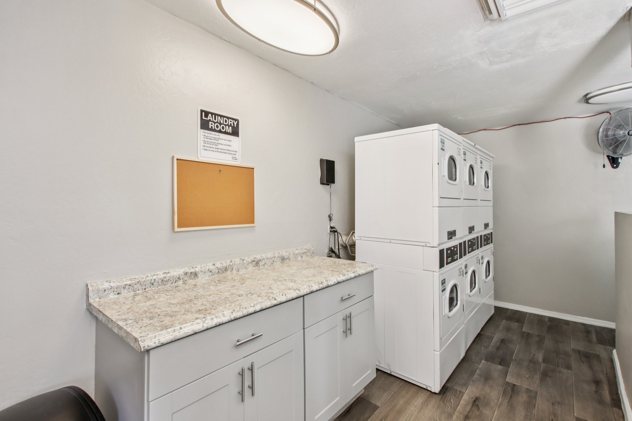 A well-lit laundry room featuring a granite countertop with cabinets underneath. Along the wall, there are stacked washers and dryers, and a bulletin board is mounted nearby. The flooring is a modern, dark wood design, and a fan is visible on the wall.