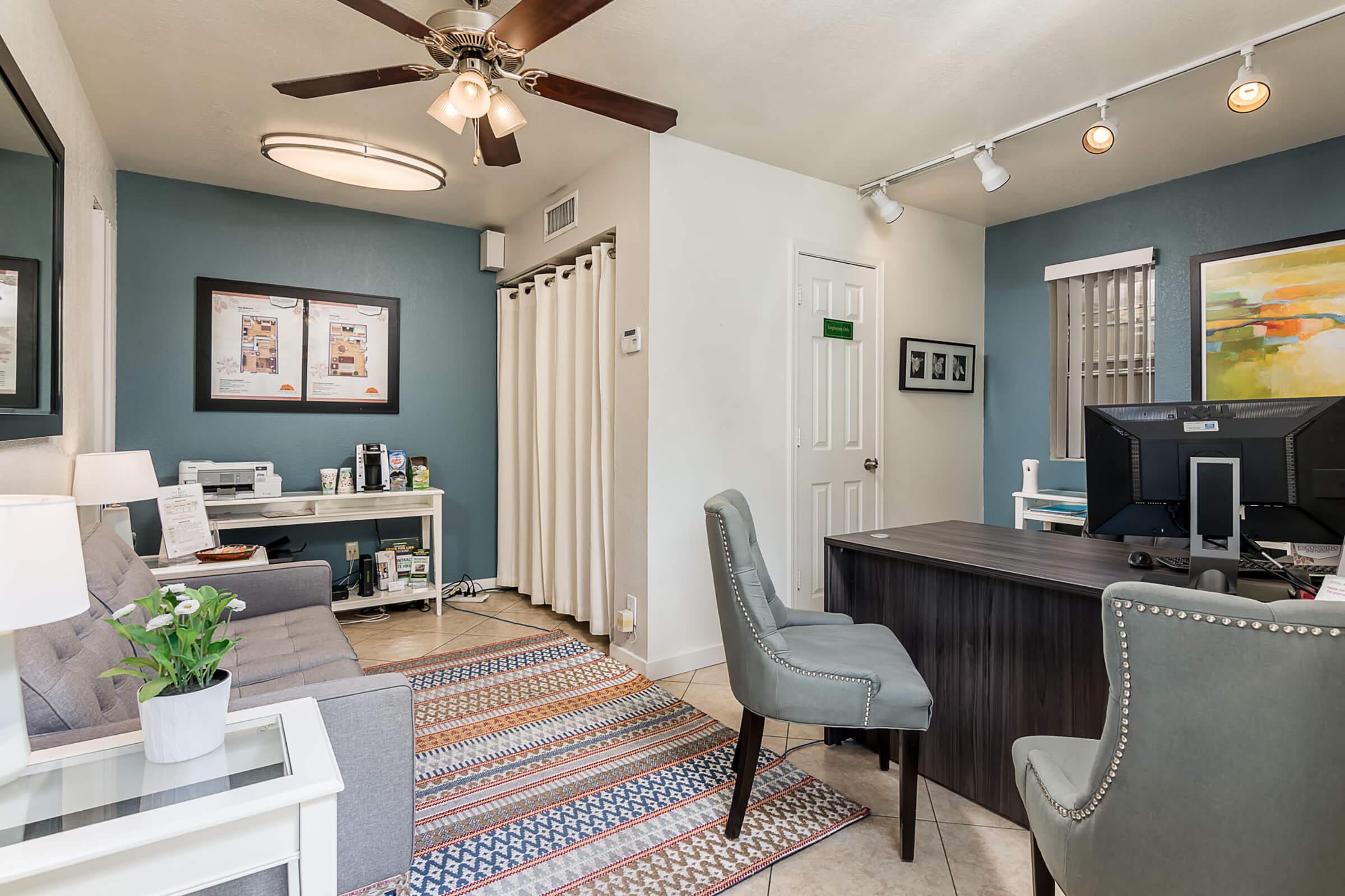 A modern office interior featuring a gray sofa, a desk with a computer, and a decorative rug. The walls are painted in blue and white, adorned with framed pictures. A ceiling fan, track lighting, and curtains add to the inviting atmosphere. The space is organized and well-lit, creating a professional environment.