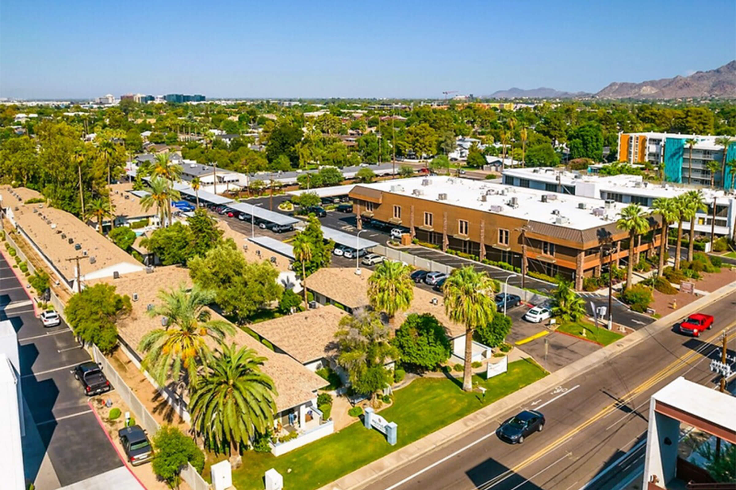 Aerial view of a suburban area featuring low-rise buildings, palm trees, and well-maintained landscaping. In the foreground, a cluster of single-story homes with textured roofs, while in the background, taller buildings are visible along a clear, blue sky. Streets are lined with cars and greenery.