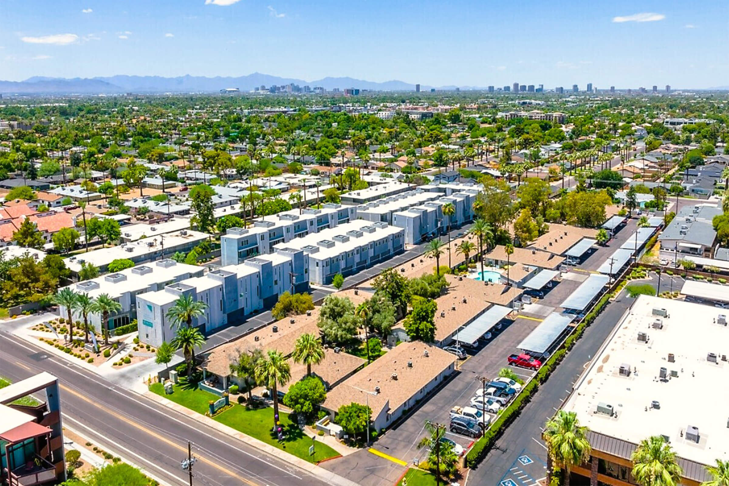 Aerial view of a suburban neighborhood featuring rows of houses and apartment buildings. The scene includes palm trees, a parking area, and some greenery. In the background, a range of mountains is visible under a clear blue sky, along with a distant urban skyline.