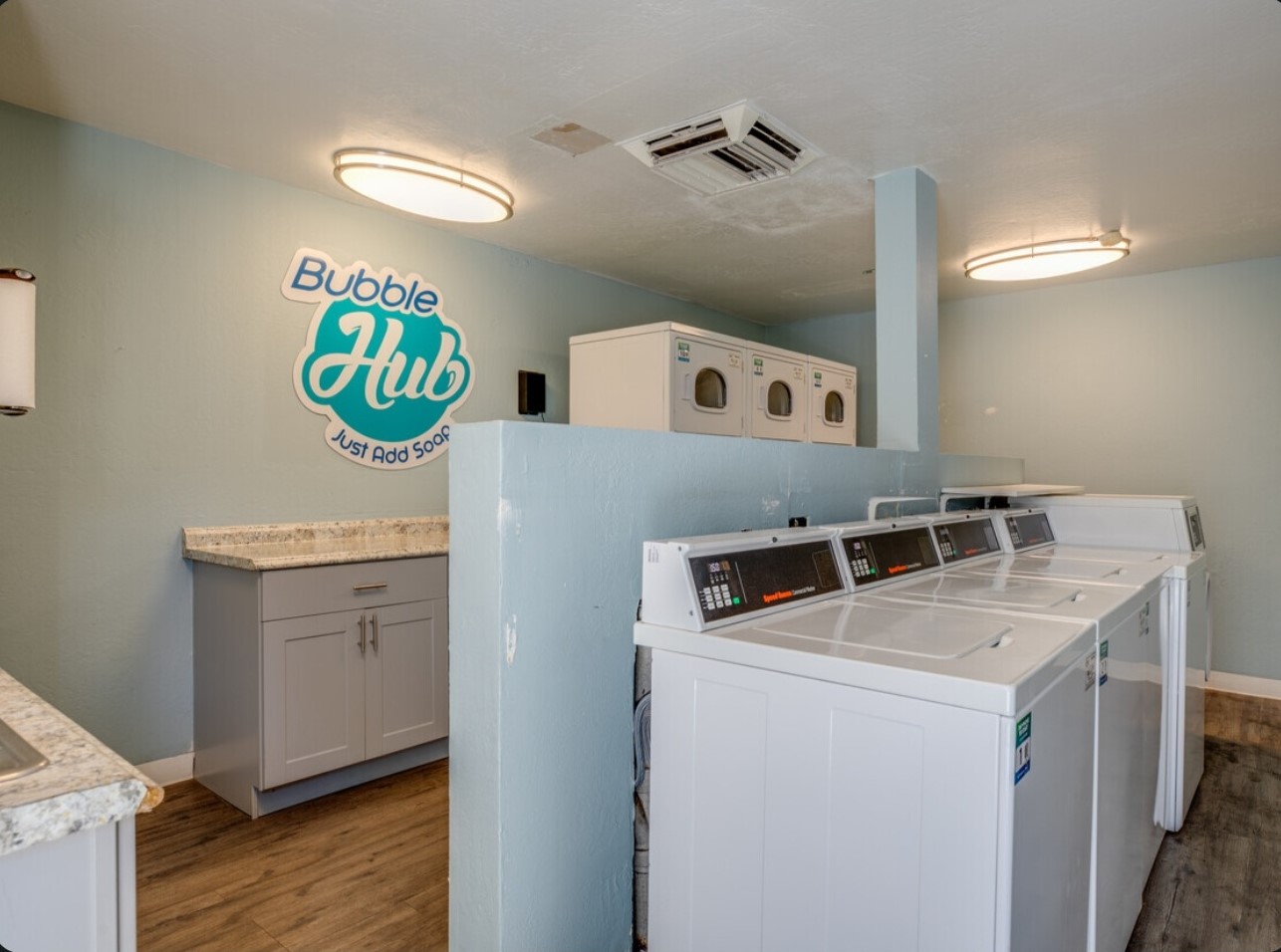 A spacious laundry room featuring a modern design with multiple washing machines and dryers. The walls are painted a light blue, and there’s a decorative sign that reads "Bubble Hub Just Add Soap." The countertop is made of granite, and overhead lighting provides a bright atmosphere.