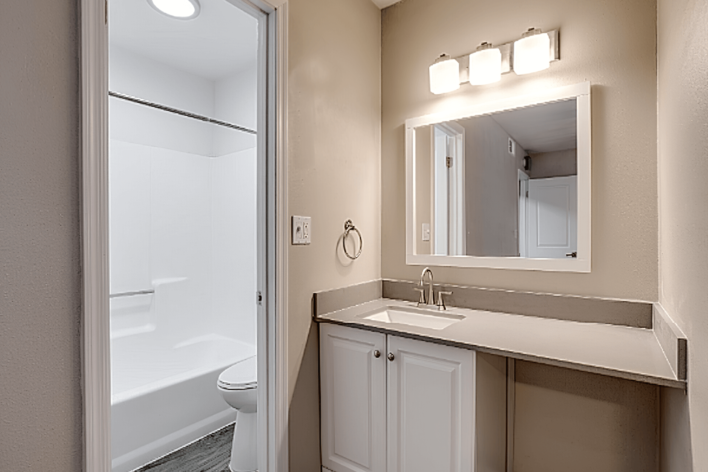 A modern bathroom featuring a white tub and shower combo, a sleek vanity with a stainless steel sink, and a large mirror above it. The walls are painted a soft beige, and there is a well-lit area with three light fixtures above the mirror. A towel ring is mounted on the wall near the sink.