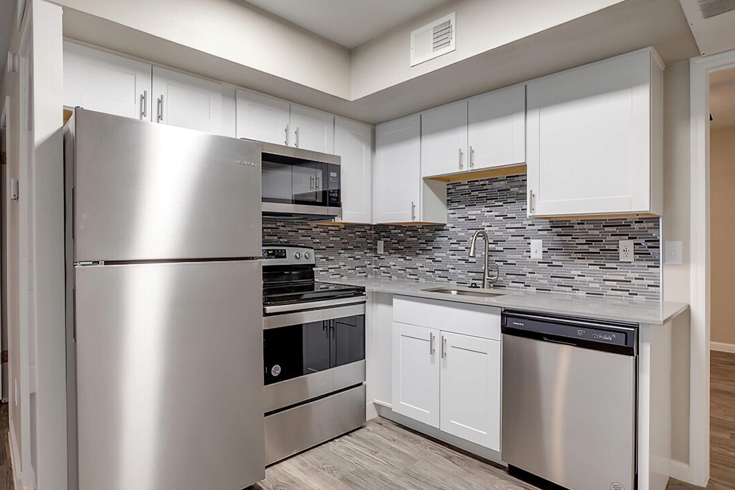 A modern kitchen featuring stainless steel appliances, including a refrigerator, oven, and dishwasher. The cabinetry is white with a tiled backsplash in gray and silver tones. Light wood flooring complements the overall design, creating a clean and contemporary look.