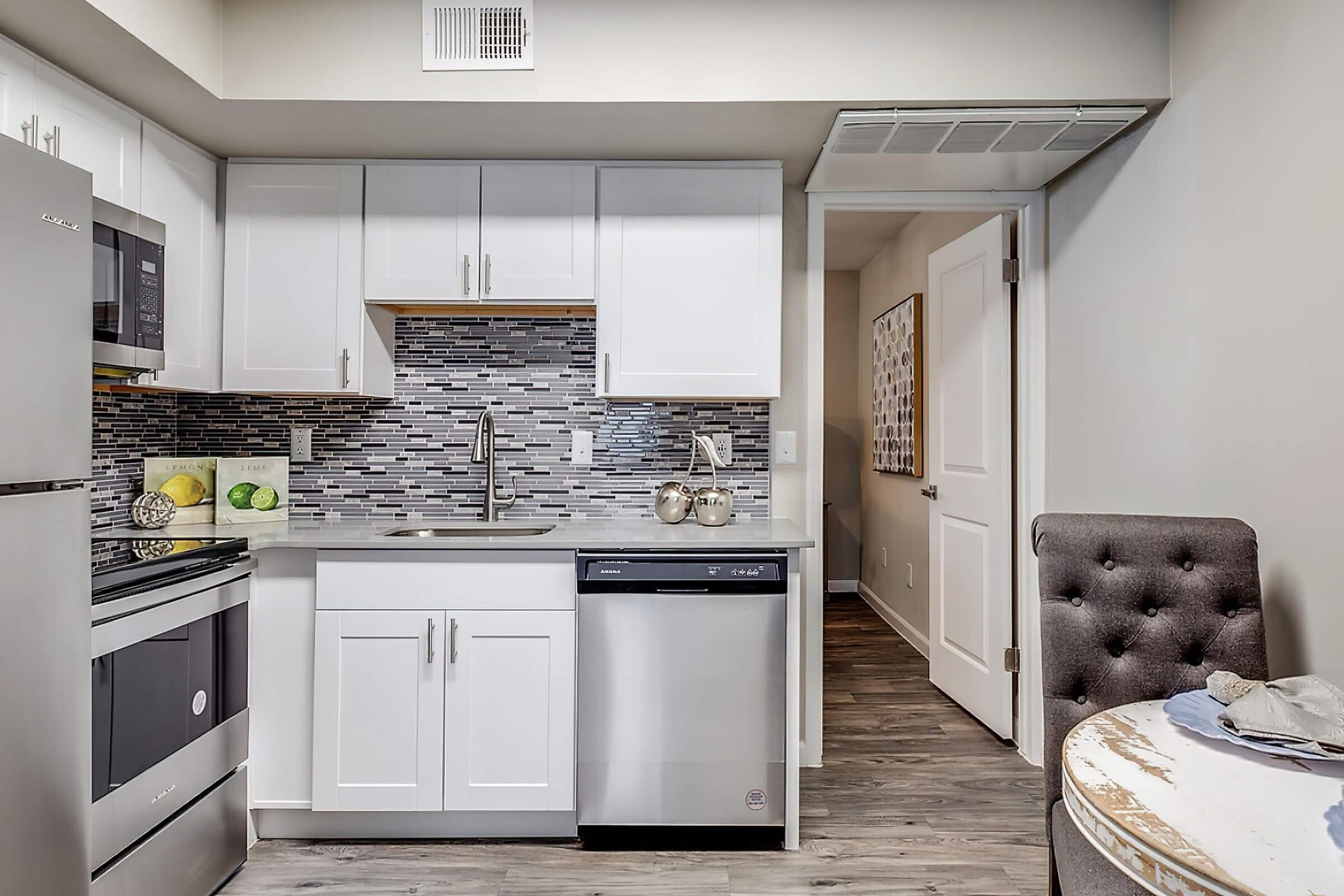 A modern kitchen featuring white cabinets, a stainless steel dishwasher, and a stove. The backsplash is made of gray and black tiles. A sink with a sleek faucet is visible, along with some colorful fruits on the counter. A dining area can be seen with a round table and a gray upholstered chair. A doorway leads to another room.