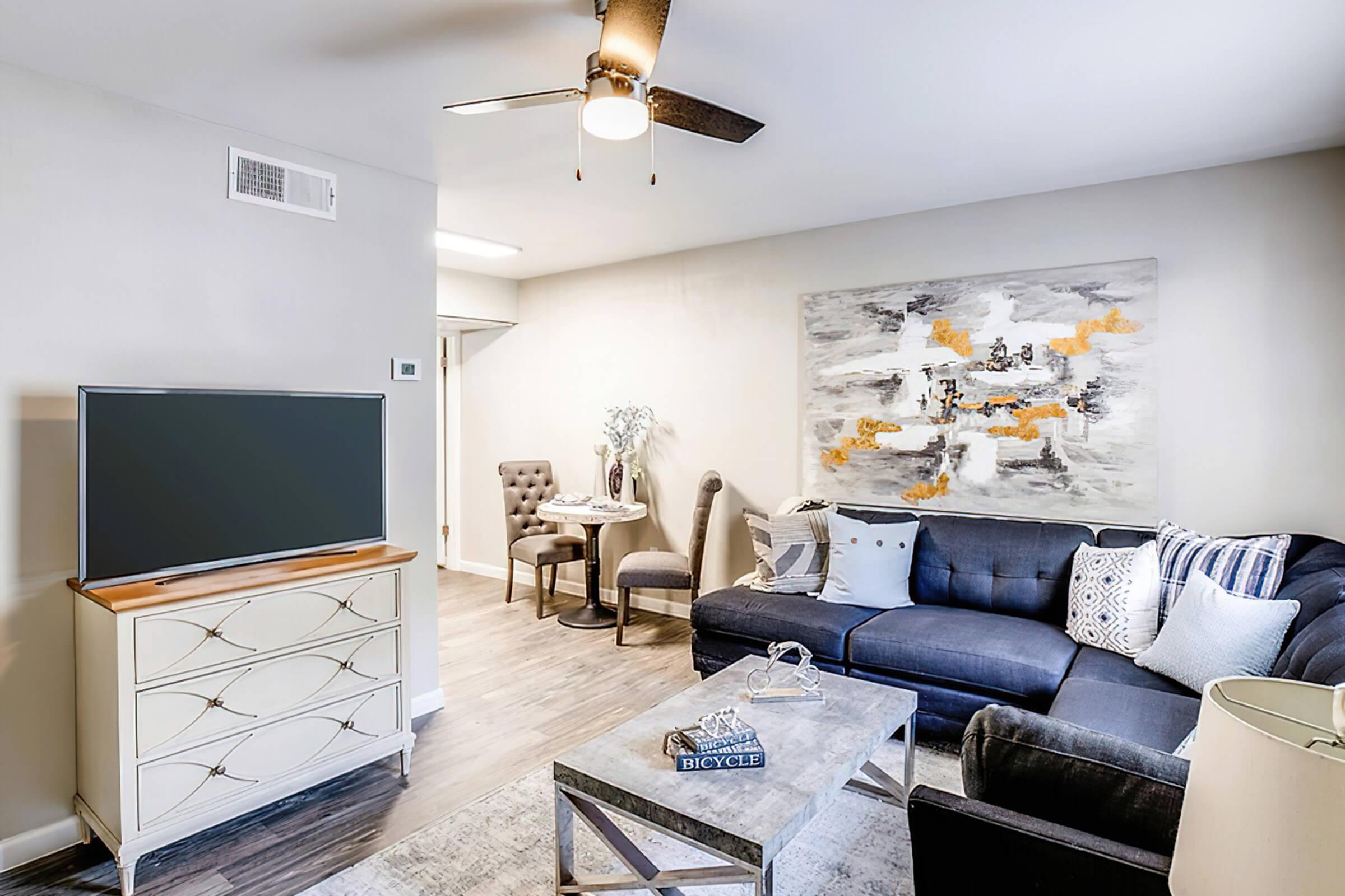 A modern living room featuring a navy blue sofa with decorative pillows, a light-colored coffee table, and a wooden TV stand. A wall-mounted fan is above, and a large abstract painting is displayed on the wall. There are two chairs and a small dining table in the background, adding to the cozy atmosphere.