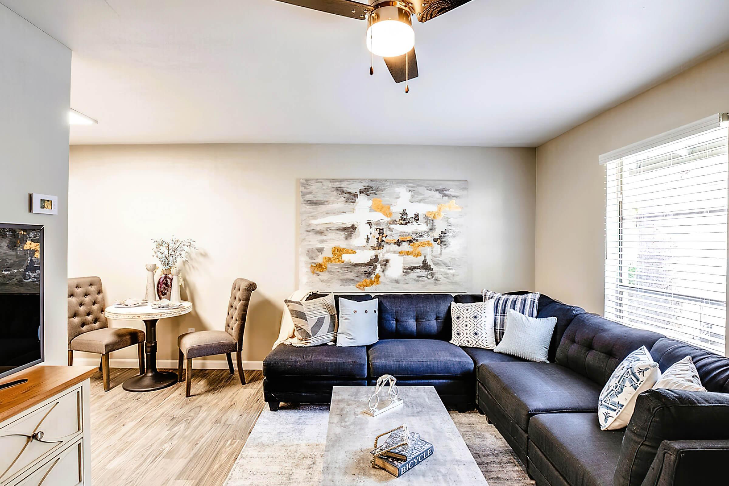 A modern living room featuring a dark blue sectional sofa adorned with decorative pillows. A glass coffee table sits in front, and a round dining table with two chairs is in the corner. A large, abstract painting with yellow and gray tones hangs on the wall, and natural light streams through a window with blinds.