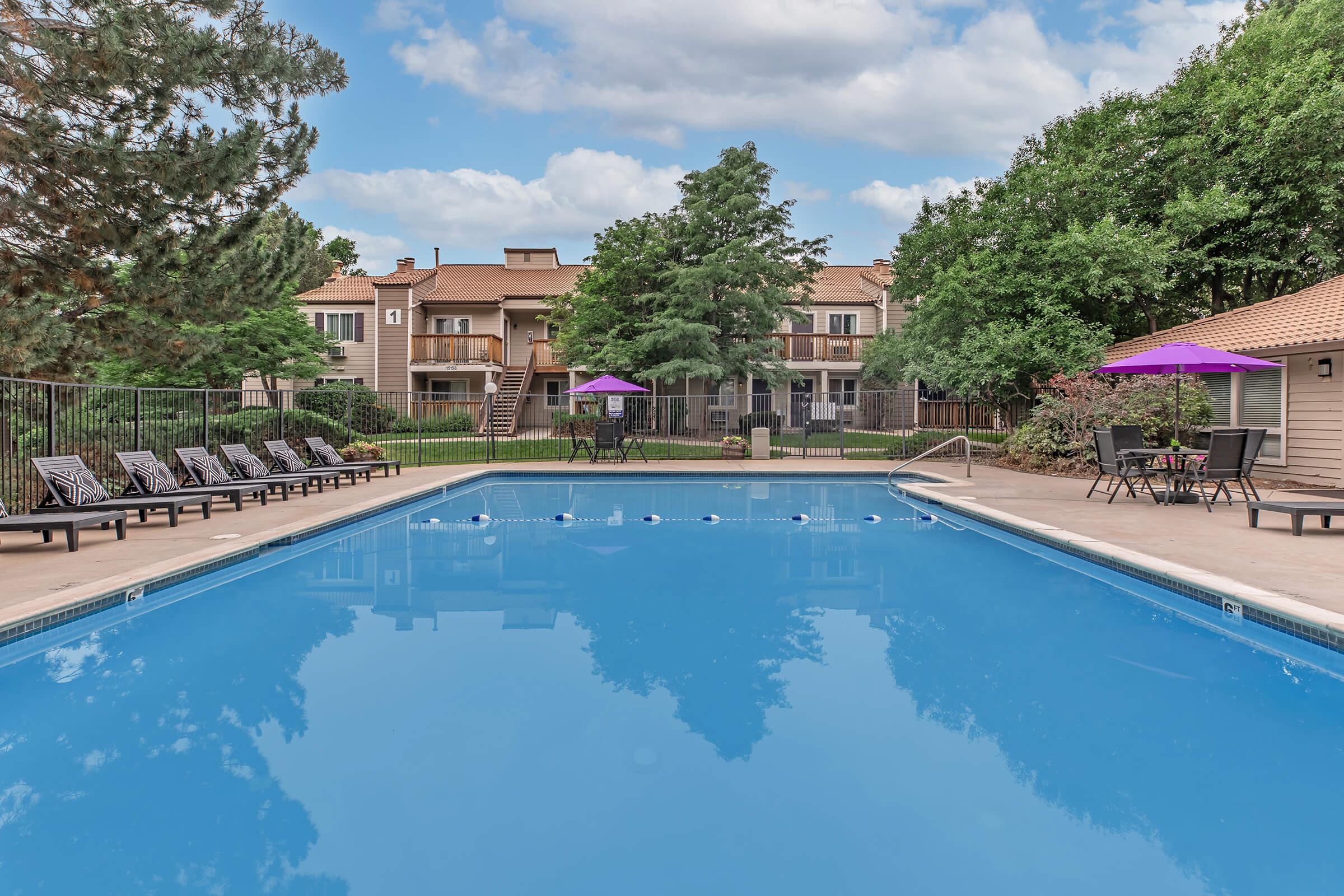 A clear blue swimming pool surrounded by lounge chairs and purple umbrellas. In the background, there are apartment buildings and lush green trees under a partly cloudy sky. The pool area is inviting and well-maintained, ideal for relaxation and leisure.