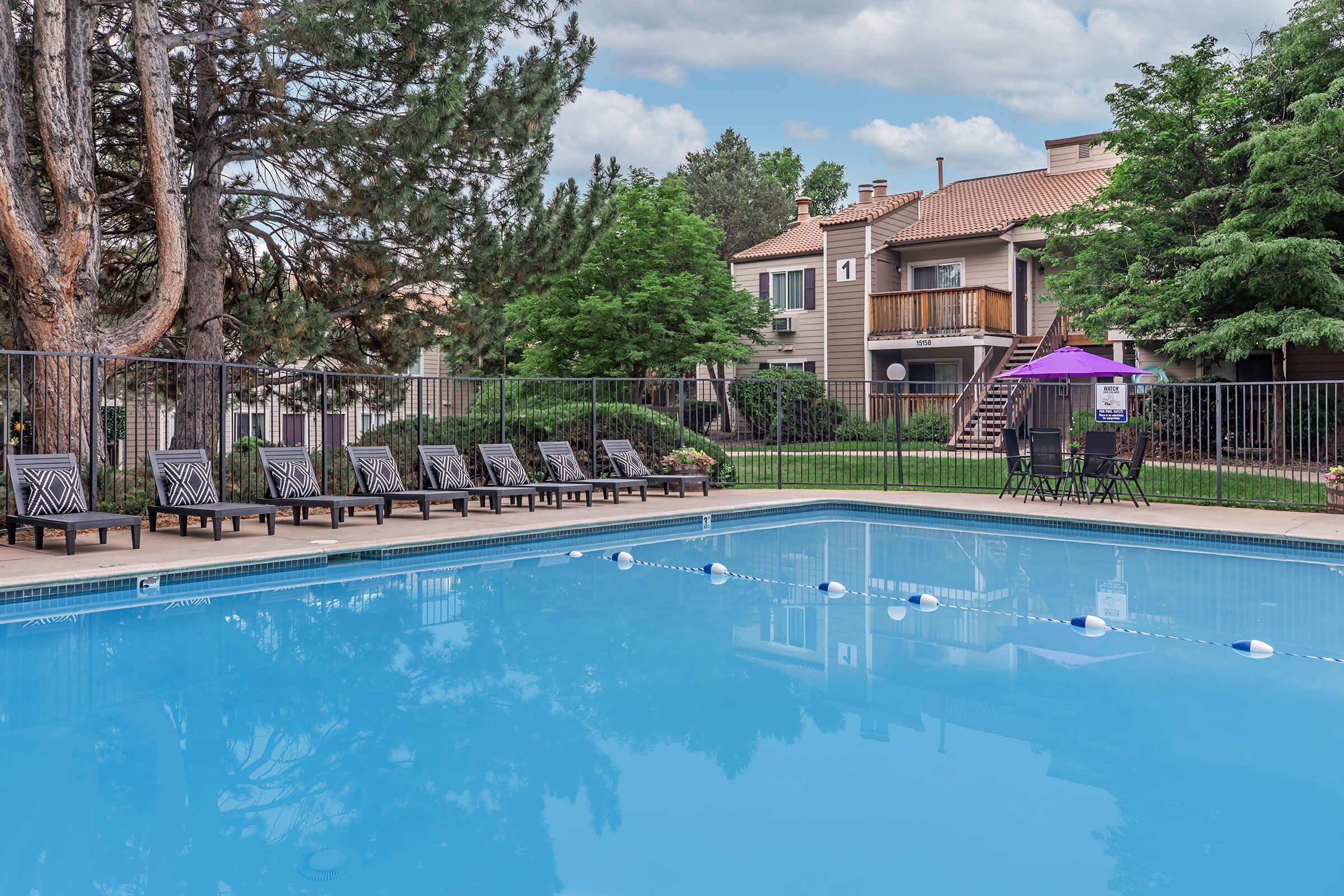 A clear blue swimming pool surrounded by lounge chairs and a fence. Lush green trees and shrubs are visible in the background, along with a few apartment buildings. The sky is partly cloudy, creating a serene outdoor setting.