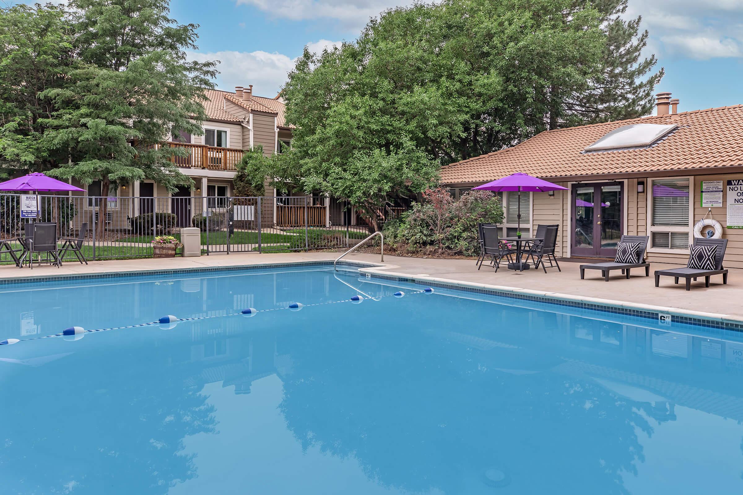 A clear blue swimming pool with lounge chairs and purple umbrellas surrounding it. In the background, there are apartment buildings partially obscured by trees and lush greenery. The scene is bright and inviting, suggesting a recreational area for relaxation and leisure.
