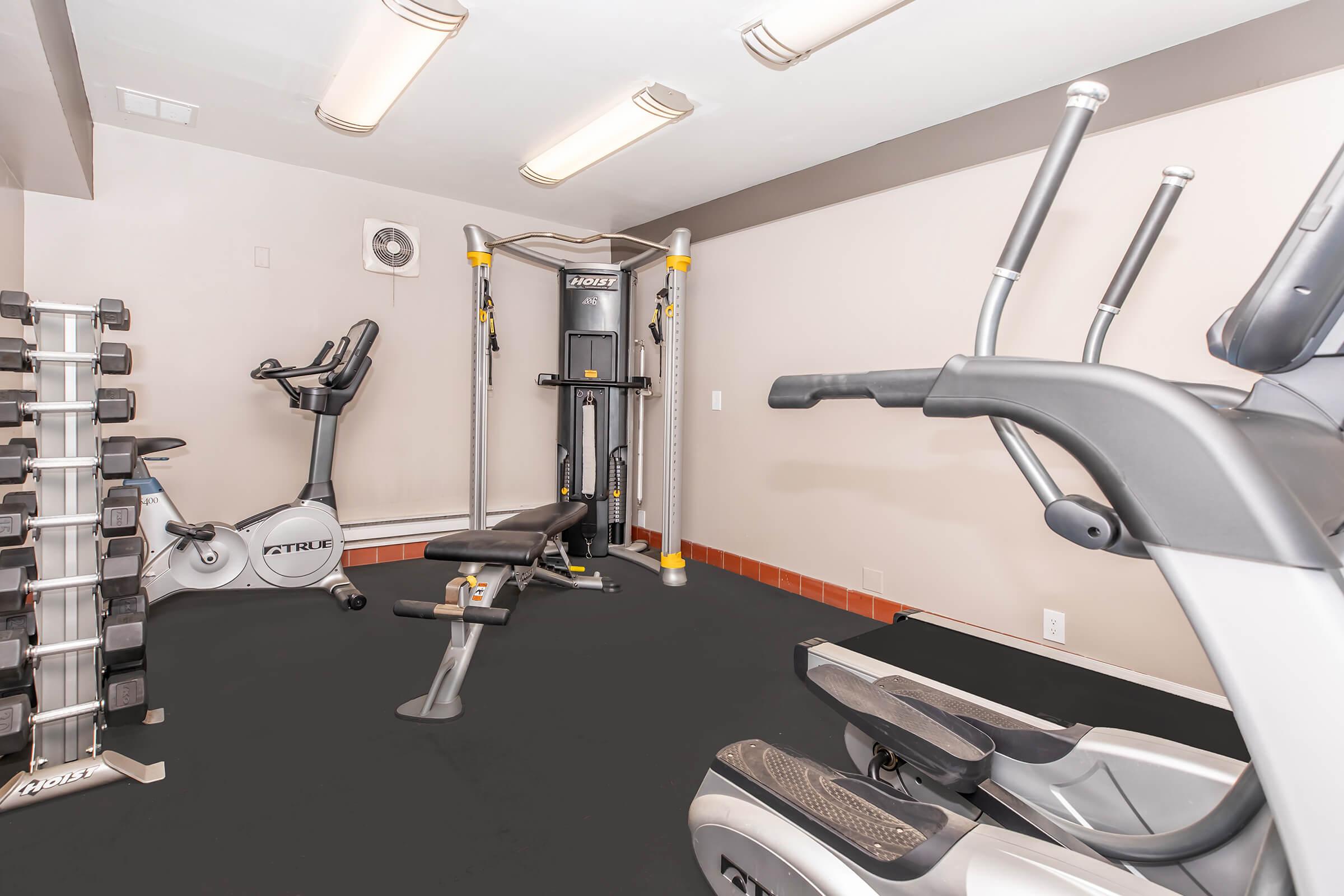 Interior view of a home gym featuring various exercise equipment. Includes a multi-station weight machine, an upright bike, and an elliptical machine, all situated on a black floor. The walls are painted in neutral tones, with overhead lighting providing ample illumination.