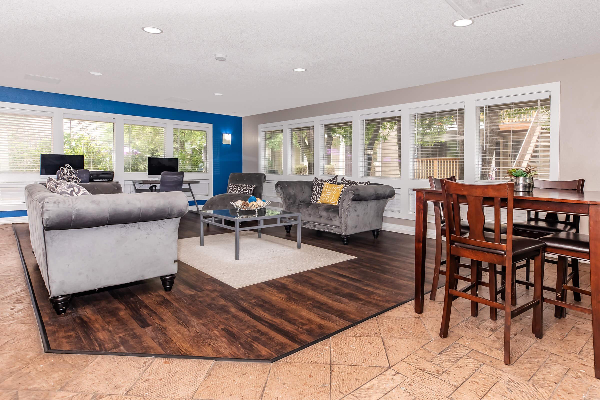 A spacious and modern living area featuring two gray sofas, a glass coffee table, and a small dining set. The room is well-lit with large windows that provide a view of the outdoors, and there is a blue accent wall. Two computers are visible in the background, adding to the modern aesthetic.