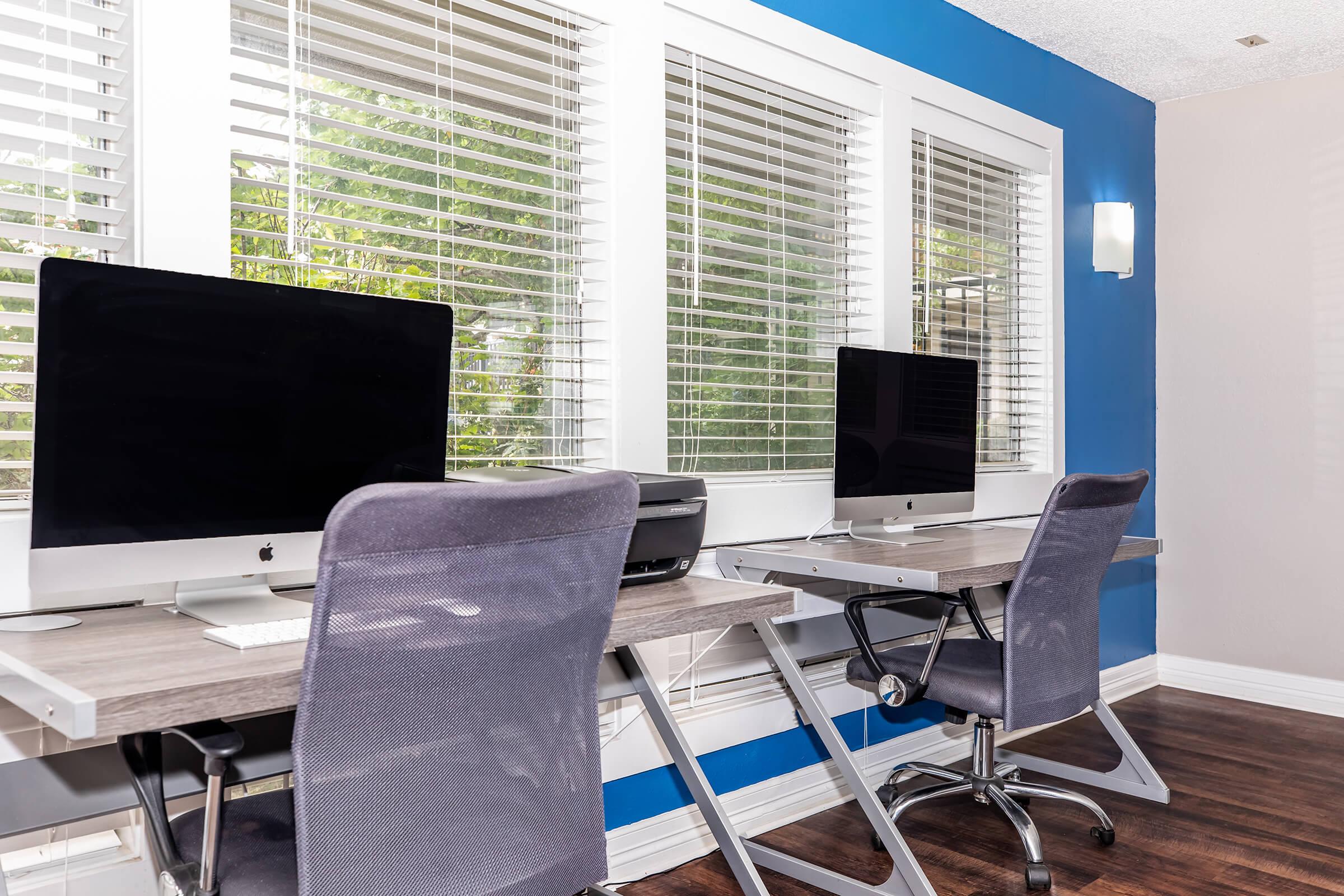 Two modern workstations with sleek design, featuring desktop computers, ergonomic chairs, and a printer. They are positioned near large windows with white blinds, allowing natural light to brighten the room. The walls are painted blue, creating a contemporary and inviting workspace environment.