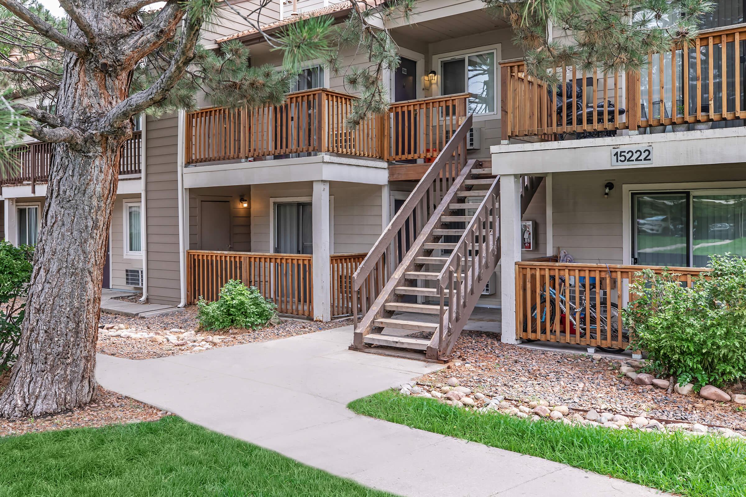 A view of an apartment complex featuring two stories with wooden balconies. A set of stairs leads to the upper level. The area includes landscaped greenery, a pathway, and a large tree nearby. The building has a welcoming appearance, with a door visible on the ground floor.