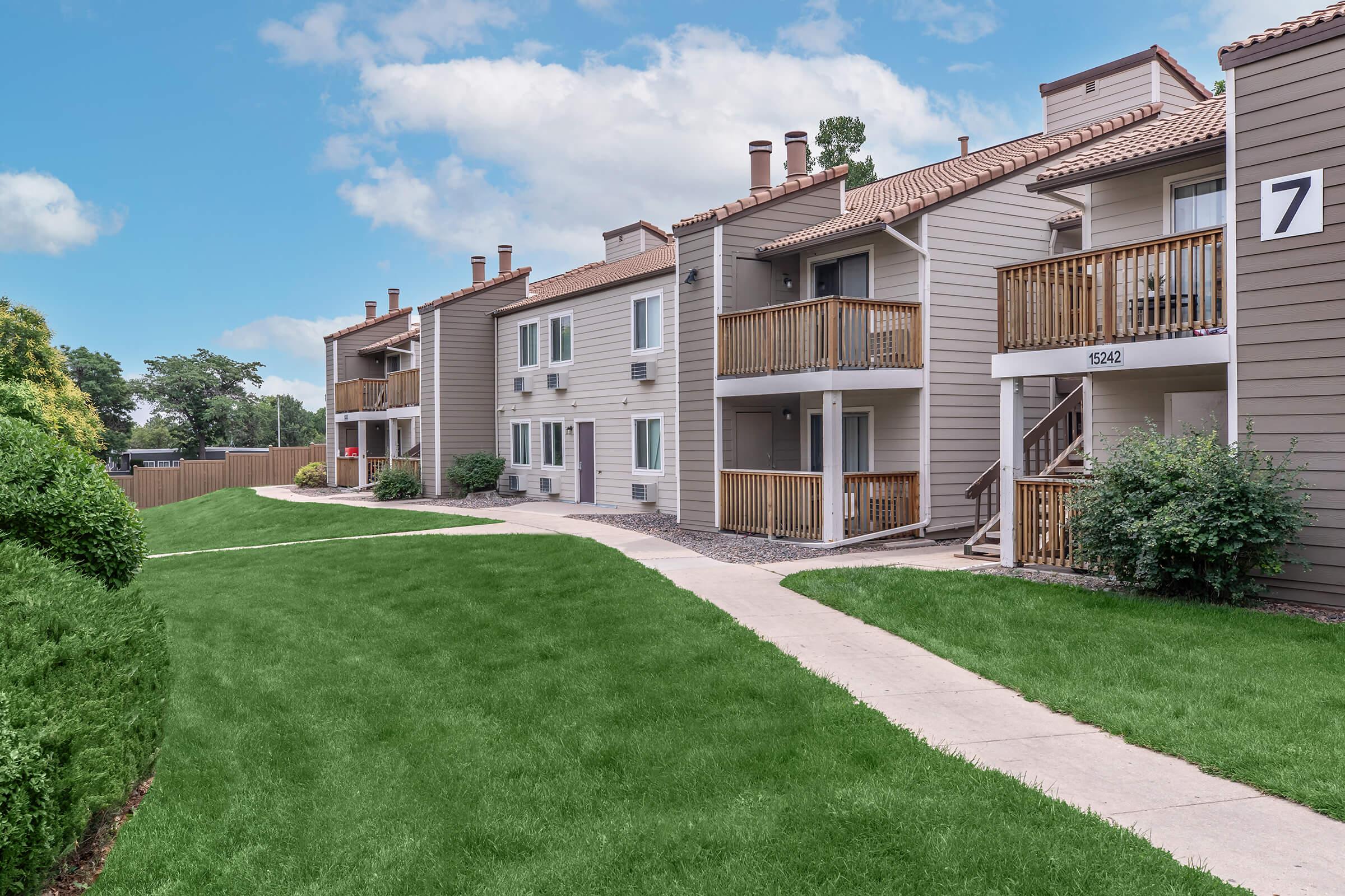 A view of a residential apartment complex featuring two buildings with balconies, surrounded by well-maintained green lawns and pathways. The sky is partly cloudy, enhancing the welcoming atmosphere of the neighborhood.