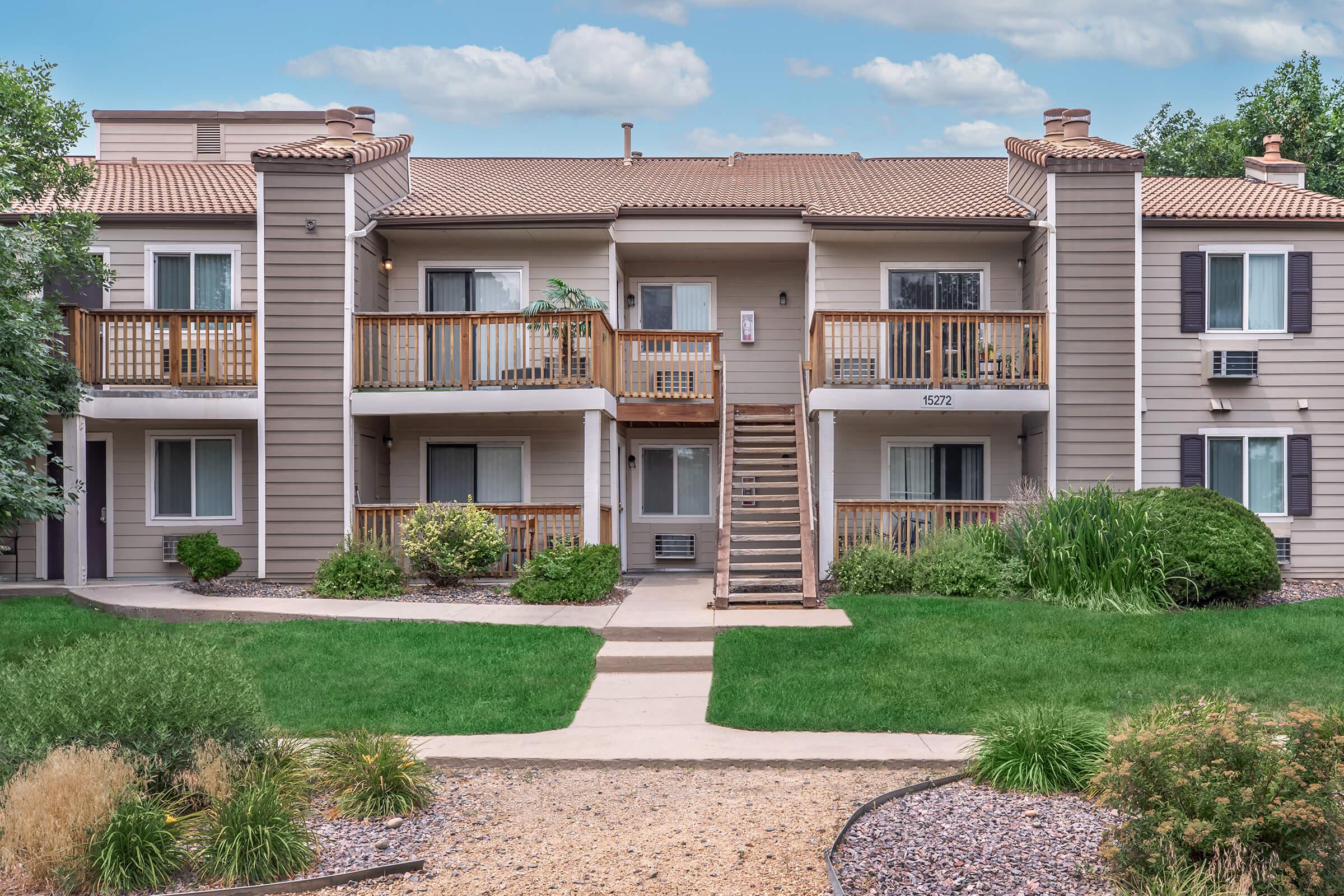 A two-story apartment building with balconies, surrounded by green grass and landscaped areas. The building features wooden railings and a staircase leading to the upper level. Shrubs and decorative stones enhance the outdoor space, creating a welcoming entrance.