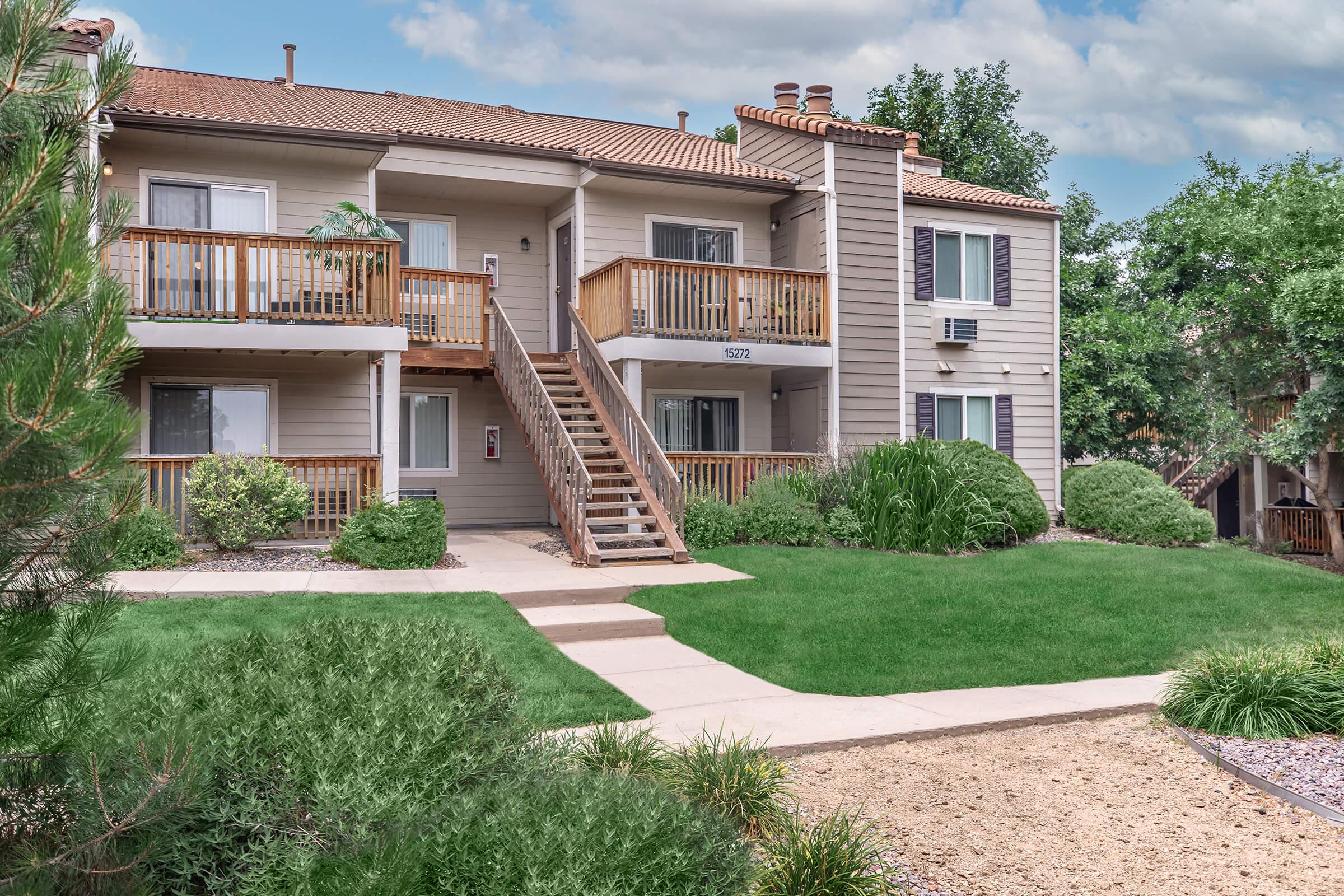 Two-story apartment building with wooden balconies and stairs, set in a green landscape. Well-maintained lawns with shrubs and small trees surround the building. The sky is partly cloudy, adding to the serene atmosphere of the residential area.
