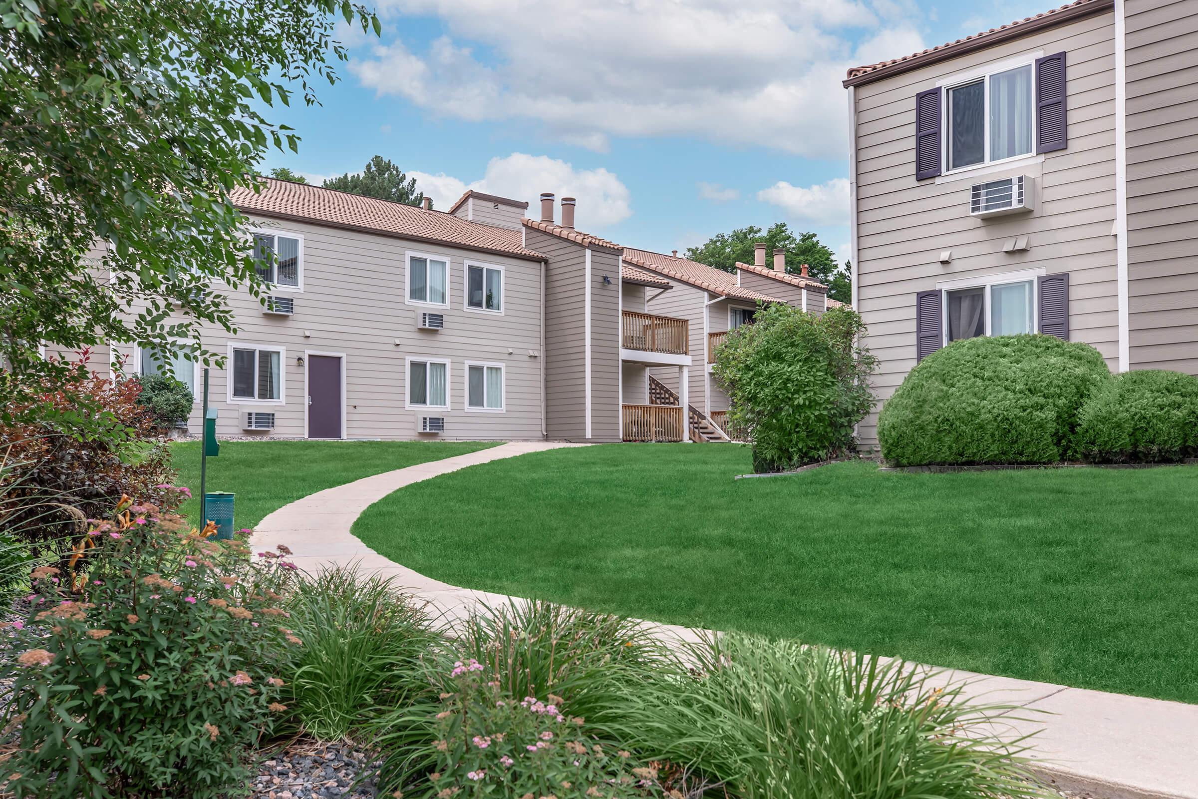 A landscaped residential area featuring two multi-unit buildings with beige siding. A curved walkway leads through a lush green lawn, surrounded by decorative shrubs and flowers. The sky is partly cloudy, adding a pleasant ambiance to the setting.